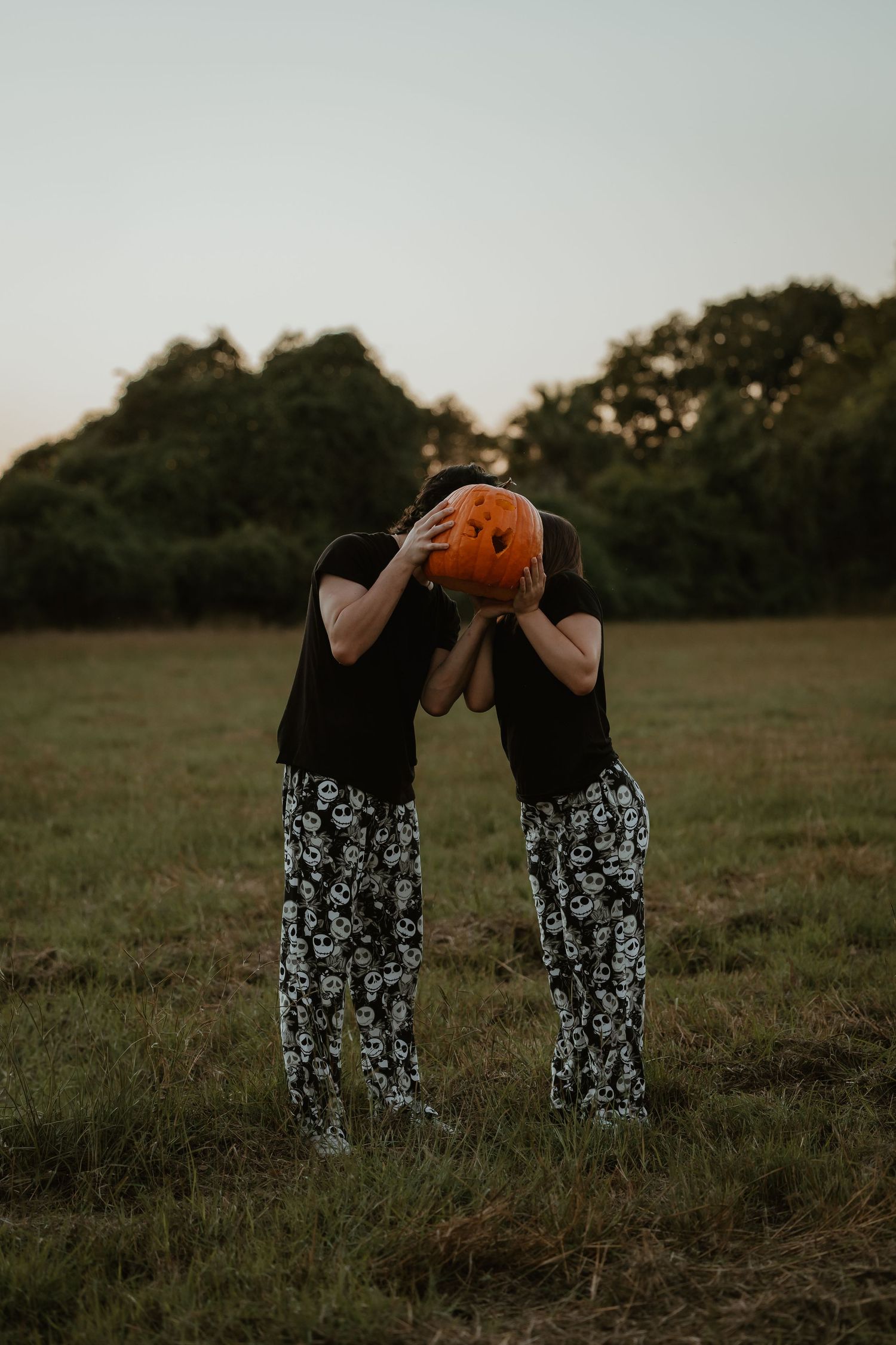 Two people in matching black tops and skull print pants hold a carved pumpkin in a grassy field at dusk.