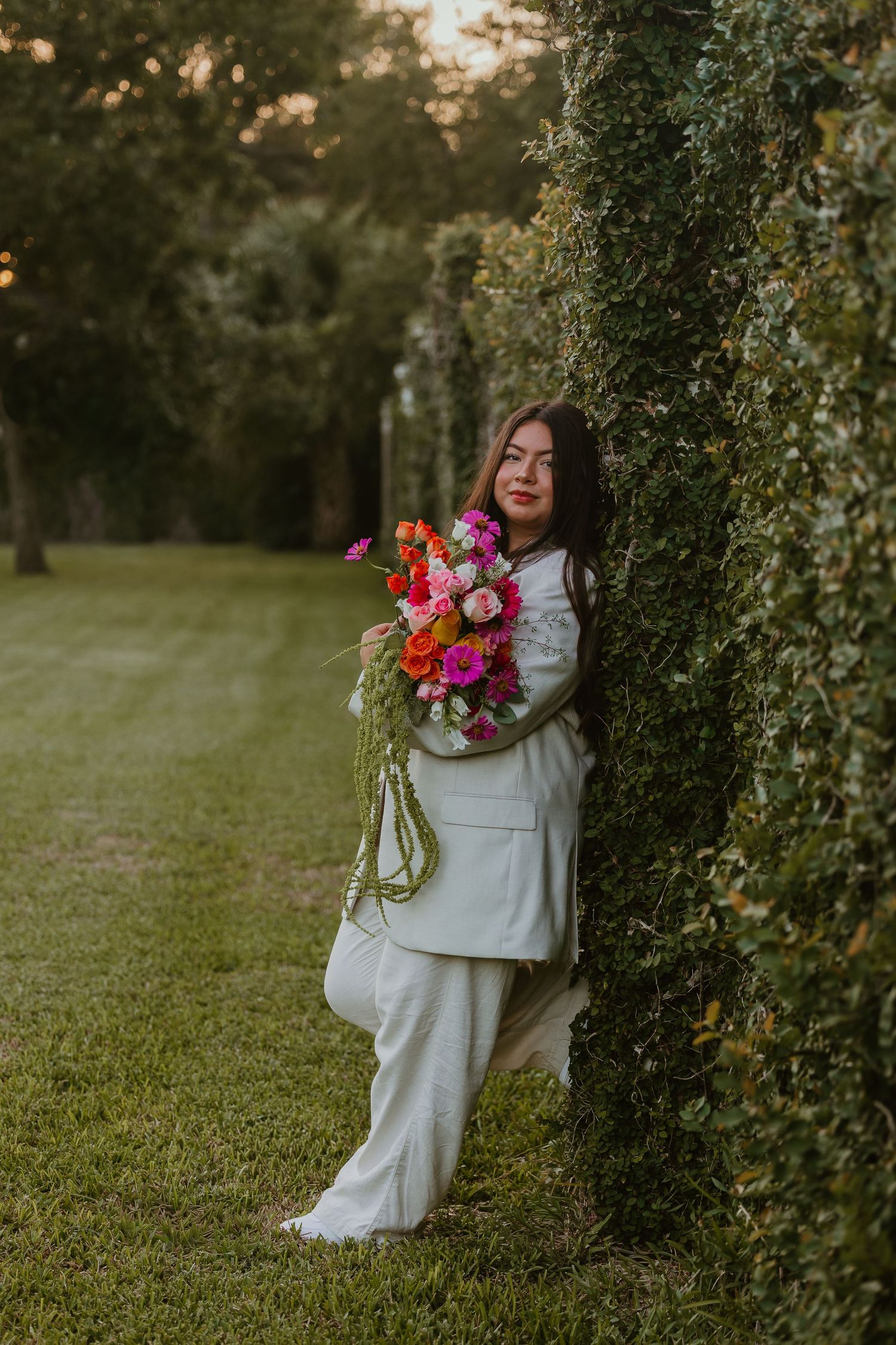 A person in a white dress holds a vibrant pink and orange bouquet while standing beside a tree at golden hour.