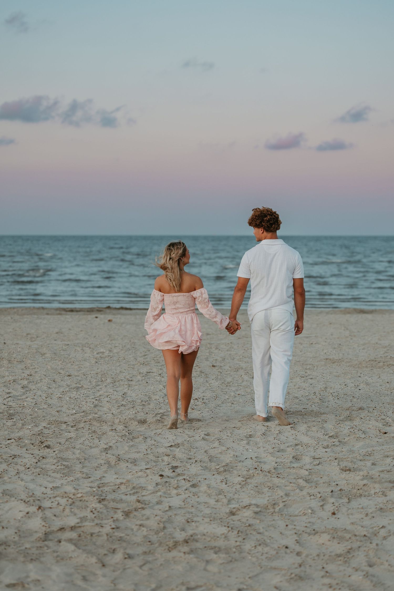 A couple walking hand in hand on a sandy beach during sunset wearing pink and white outfits.