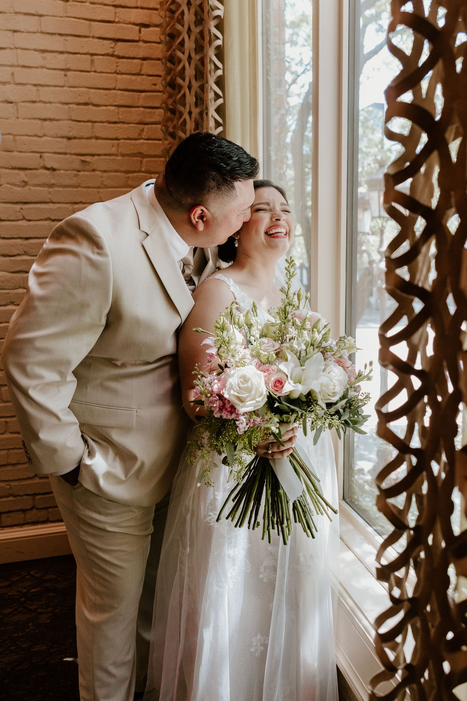 Wedding couple sharing an intimate moment by a window with sunlight and floral bouquet.