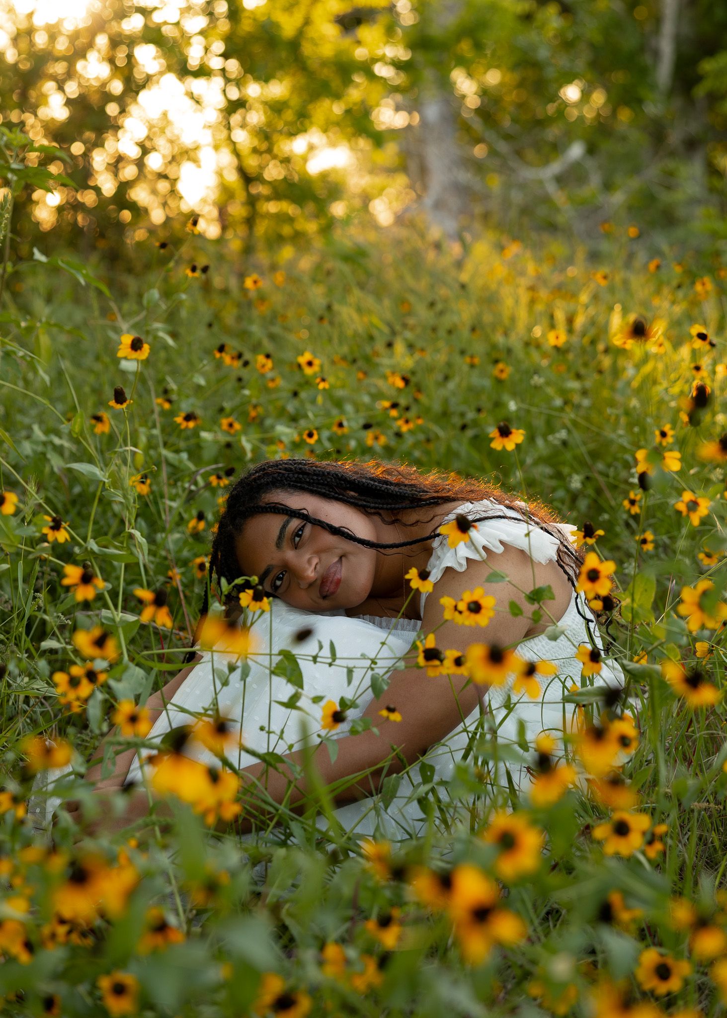 Young woman in white dress lying among yellow wildflowers in a sunlit meadow, smiling softly at the camera.