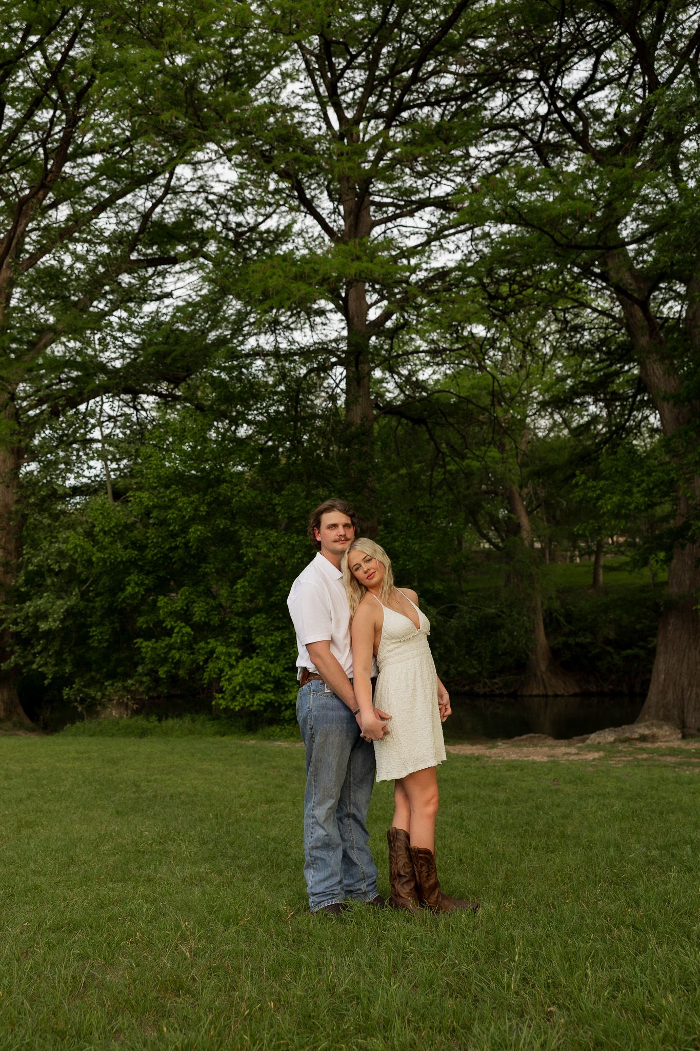 Couple embracing in a lush green park, woman in white dress and boots leans on man in white shirt and jeans.