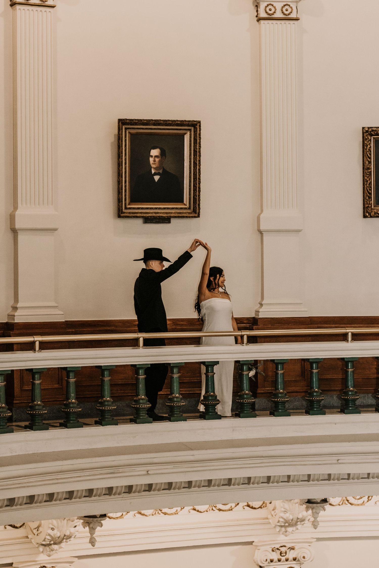 A couple poses for artistic photos in an elegant indoor venue with white columns and portrait paintings.