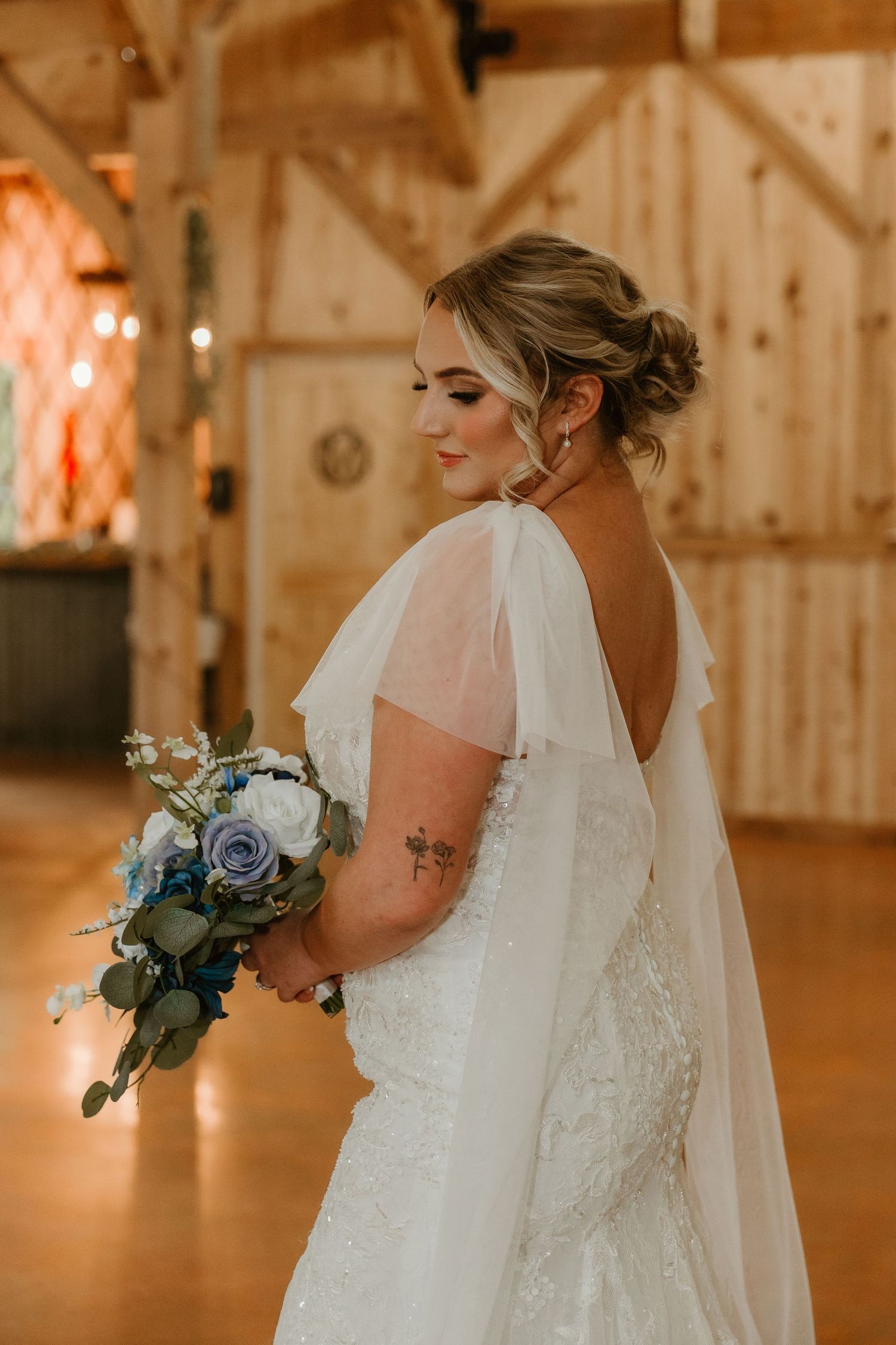 Bride in elegant white lace wedding dress with flowing veil holds blue bouquet in rustic barn venue with warm lighting.