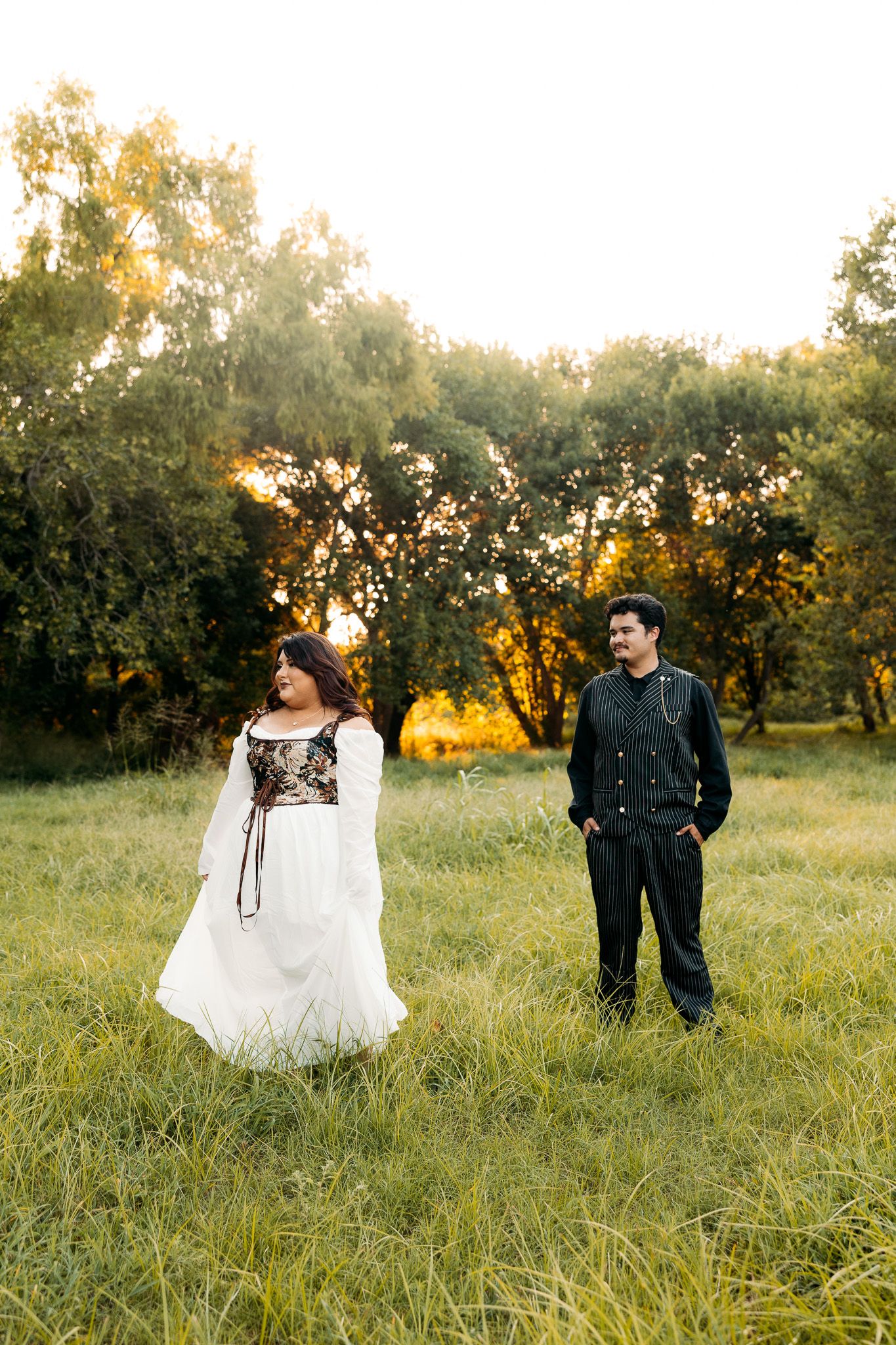 Couple standing in sunlit green meadow at golden hour, woman in white embroidered gown, man in black suit smiling.