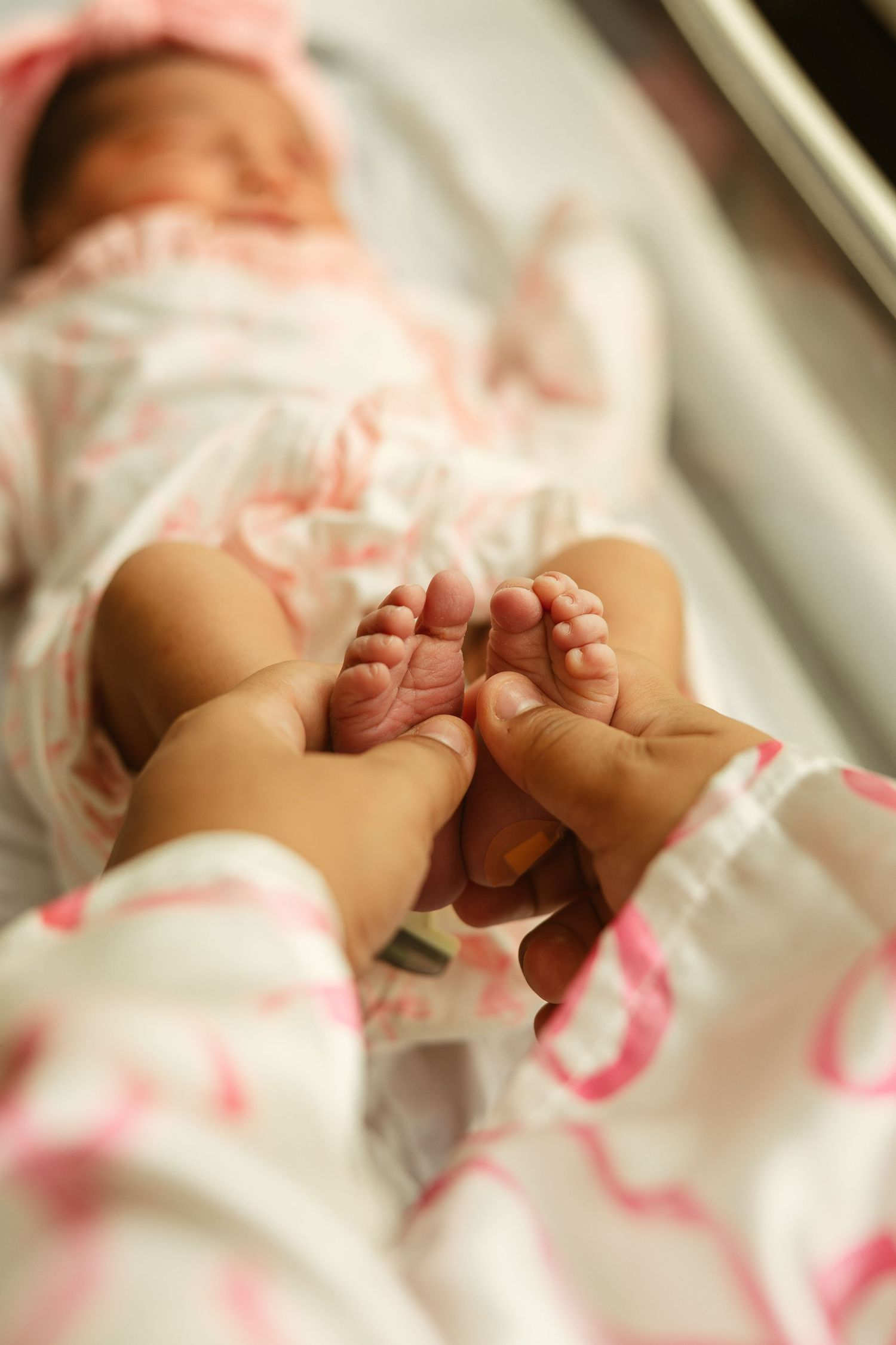 Adult hands gently holding tiny newborn baby feet while infant rests in hospital bassinet wearing pink outfit.