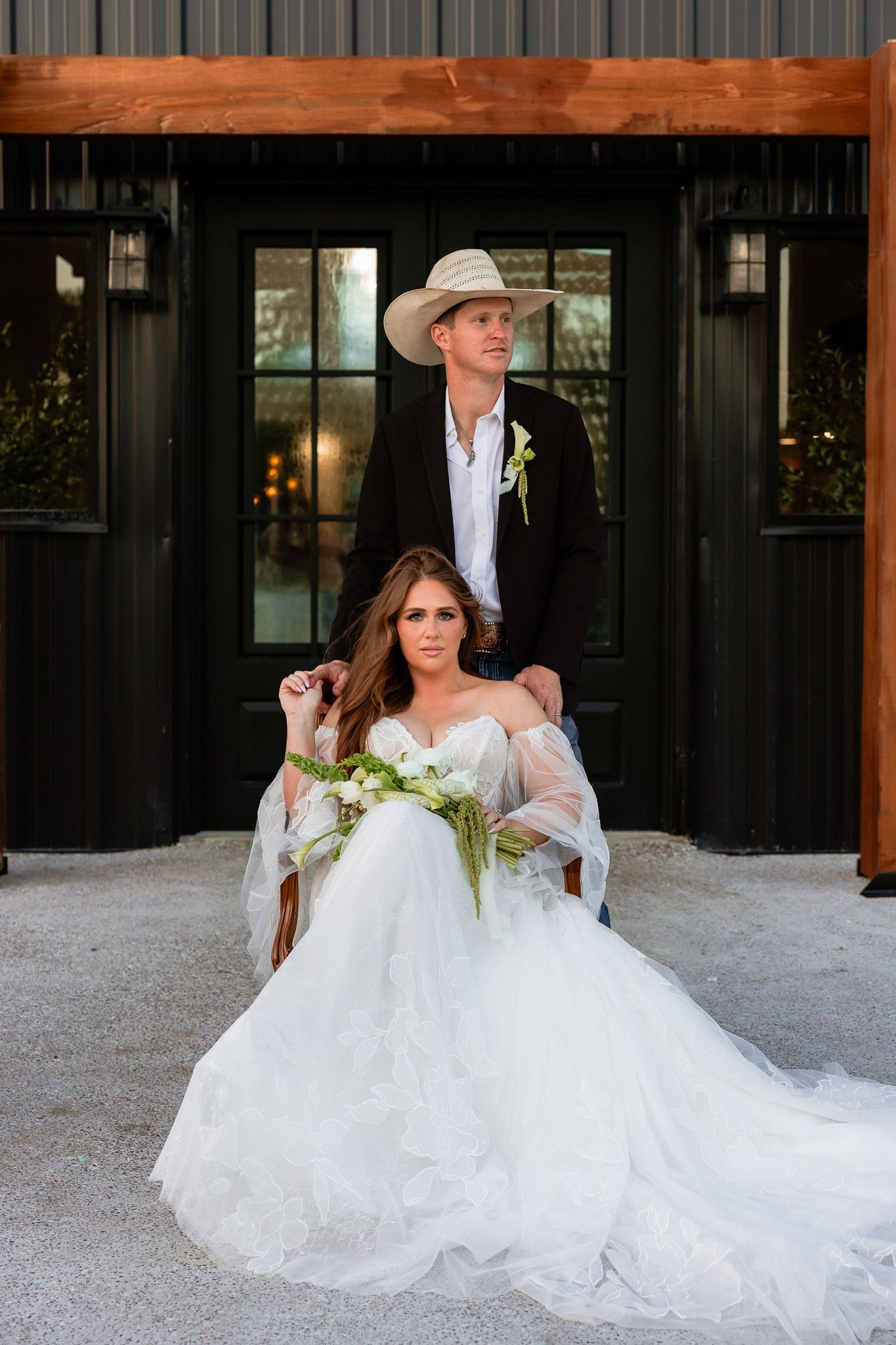Bride in white tulle gown seated holding greenery bouquet with groom in cowboy hat and black blazer standing behind her.