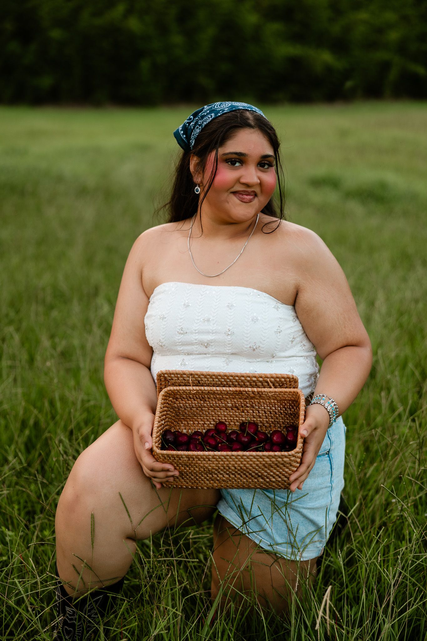 Young woman holding a wicker basket of fresh raspberries while kneeling in a green field wearing a white tube top.