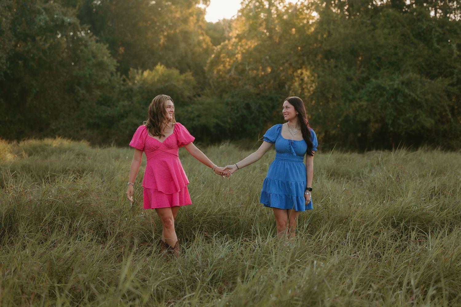 Two friends in pink and blue dresses dance playfully in a sunlit meadow at golden hour.