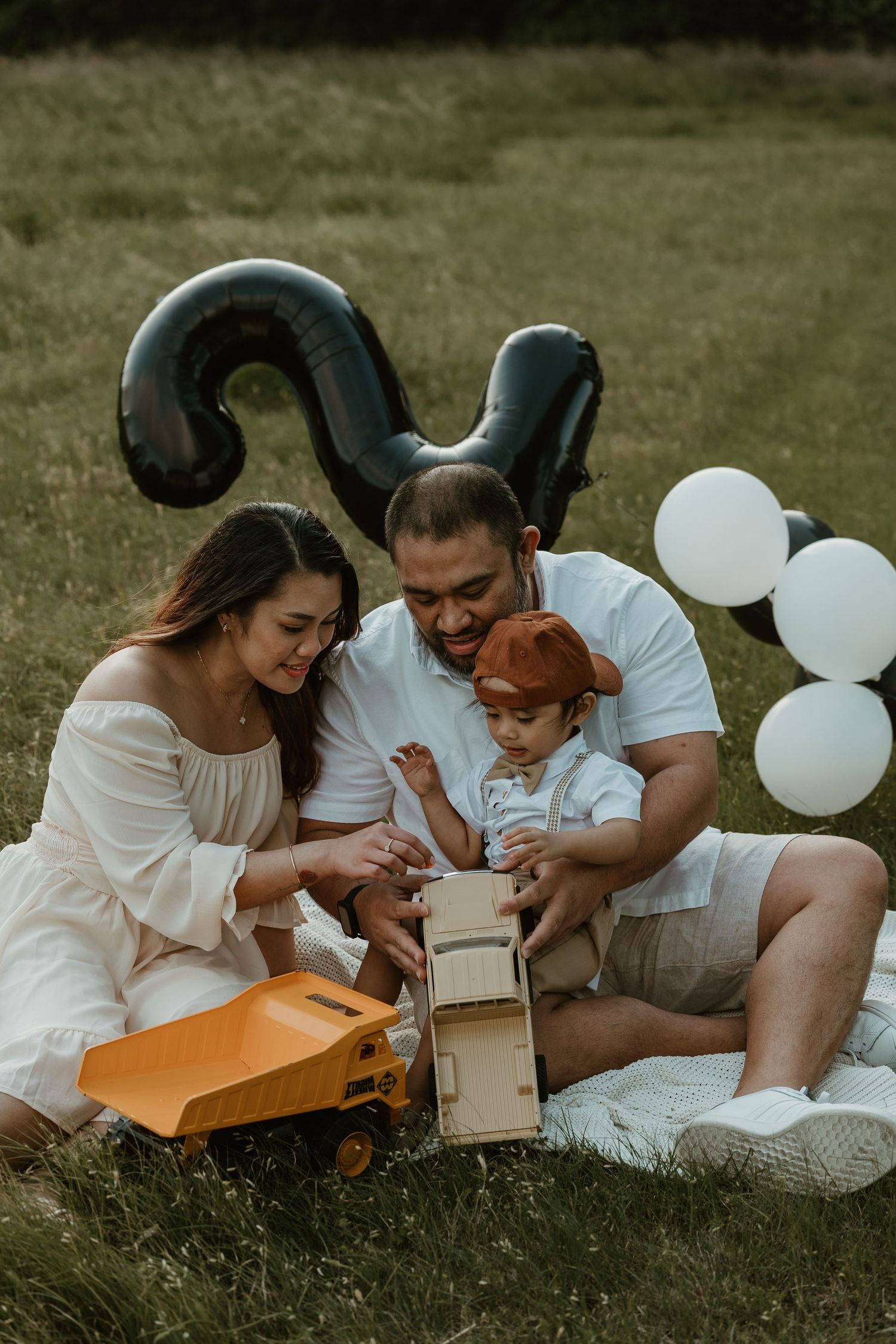 Family sitting on picnic blanket with balloons and toy truck opening packages together.