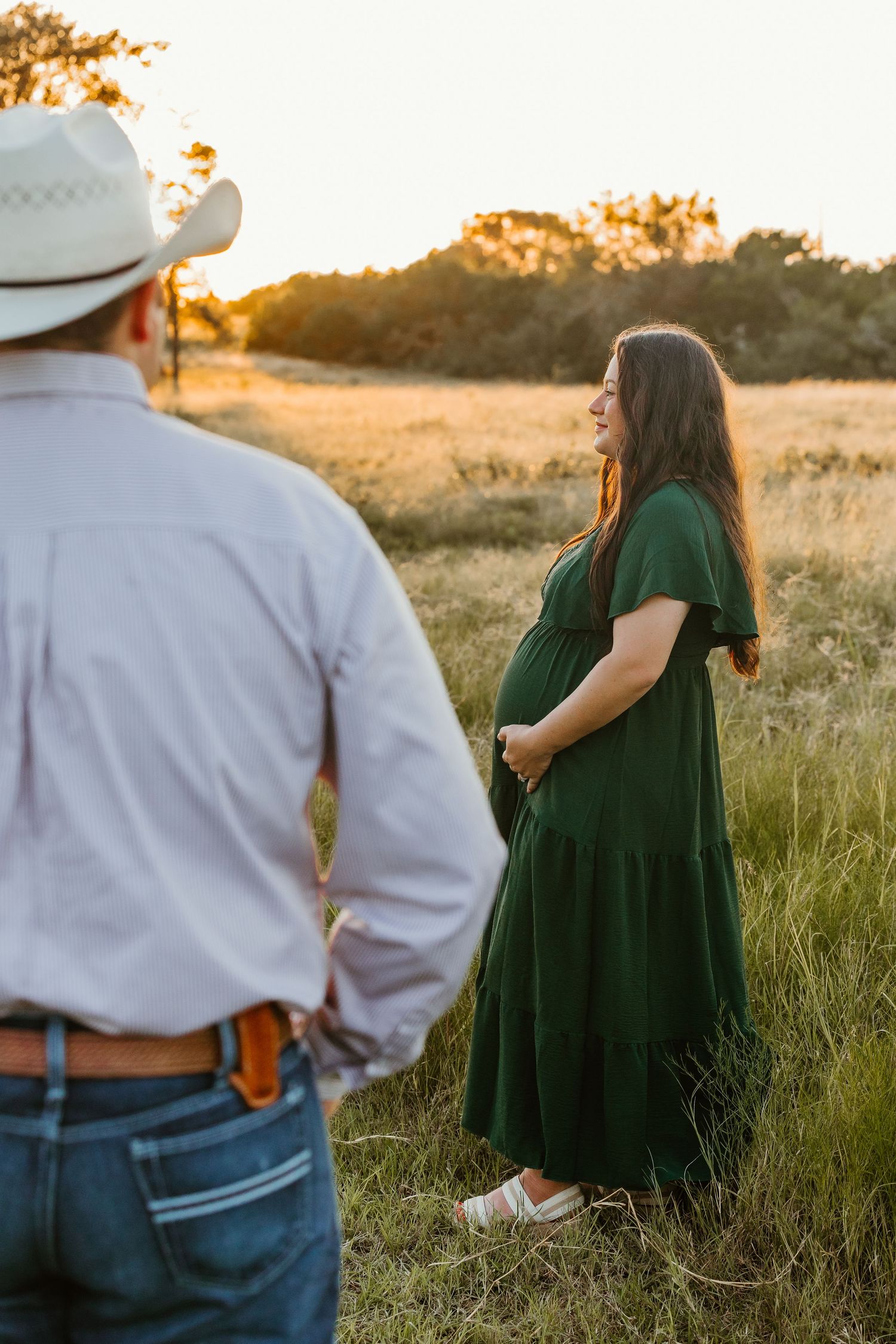 A couple stands in a sunlit field during their maternity photography session.