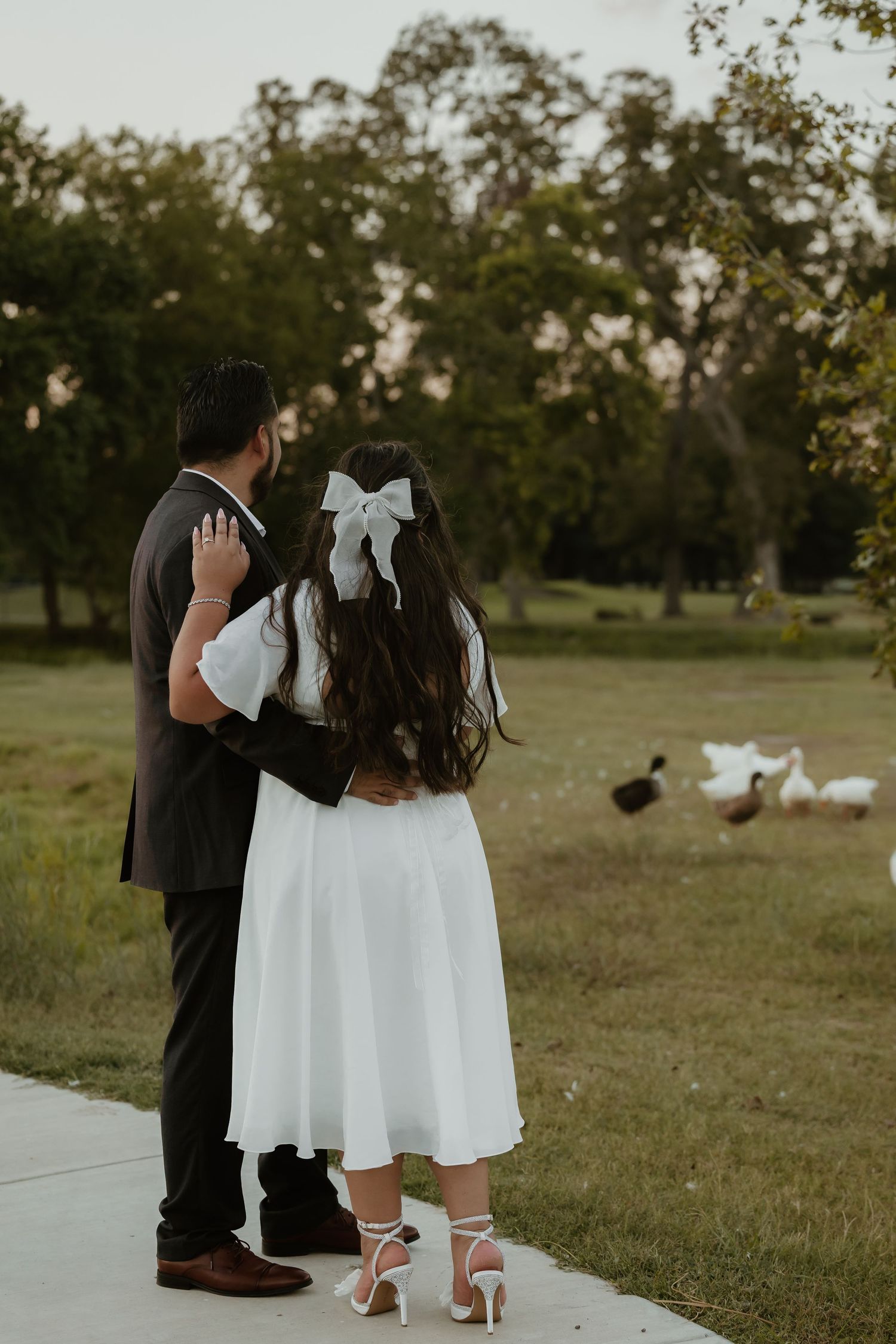 Couple in formal attire stands on pathway with ducks in background.