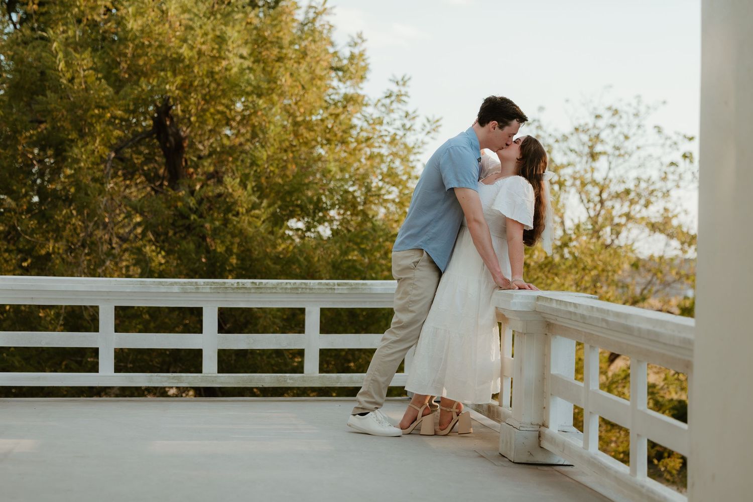 Romantic couple embraces on a white railing bridge during golden hour with autumn trees in background.