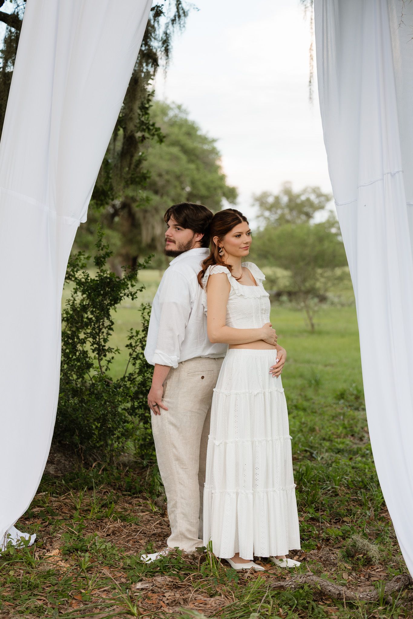 Couple in white attire standing back-to-back under draped fabric outdoors, woman cradling baby bump in lush green setting.