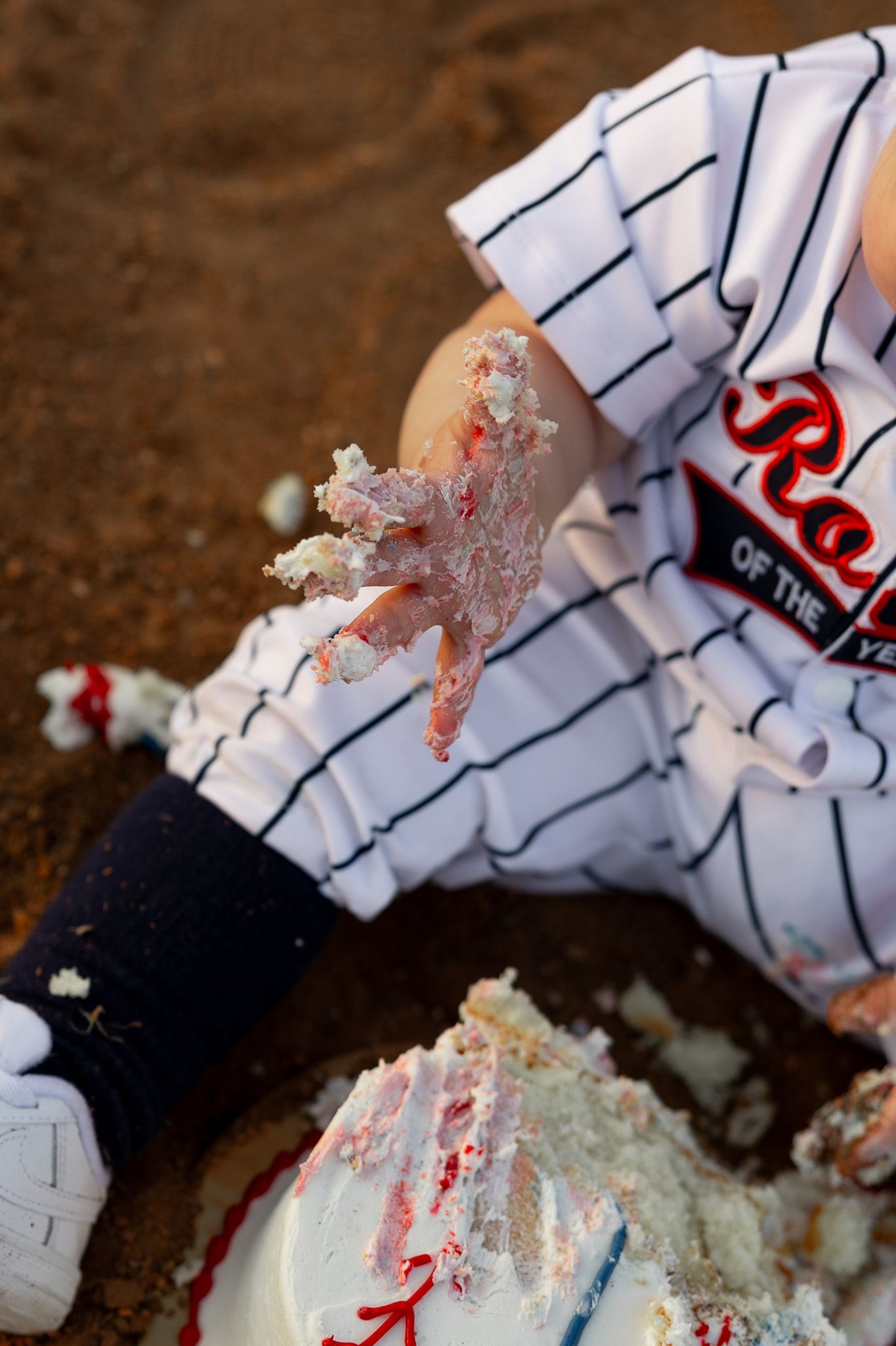 Baby in baseball uniform smashing first birthday cake on dirt field, frosting on tiny hand.