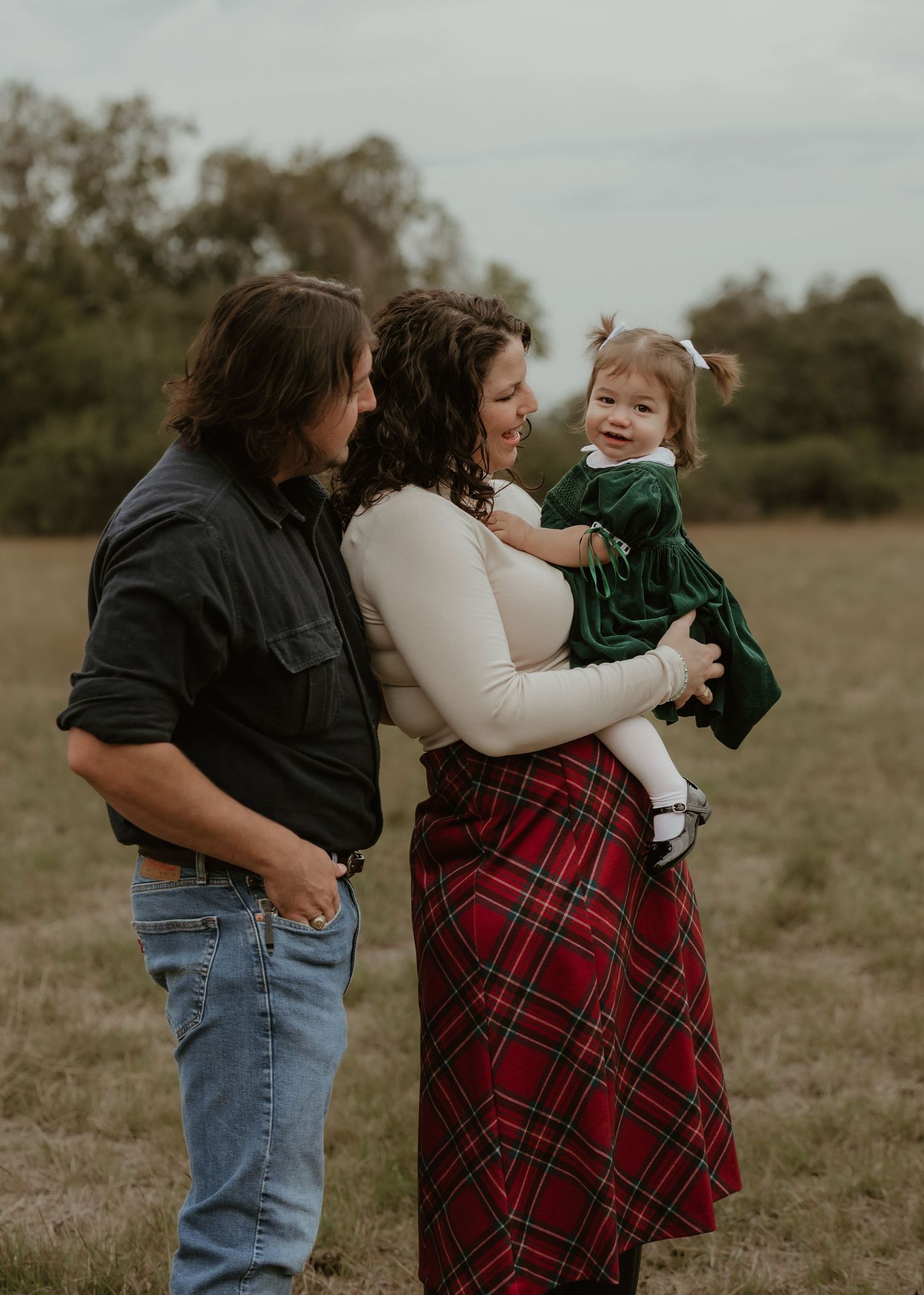 Family of three posing outdoors in a field, parents smiling at baby girl in green dress, holiday plaid skirt and black shirt.