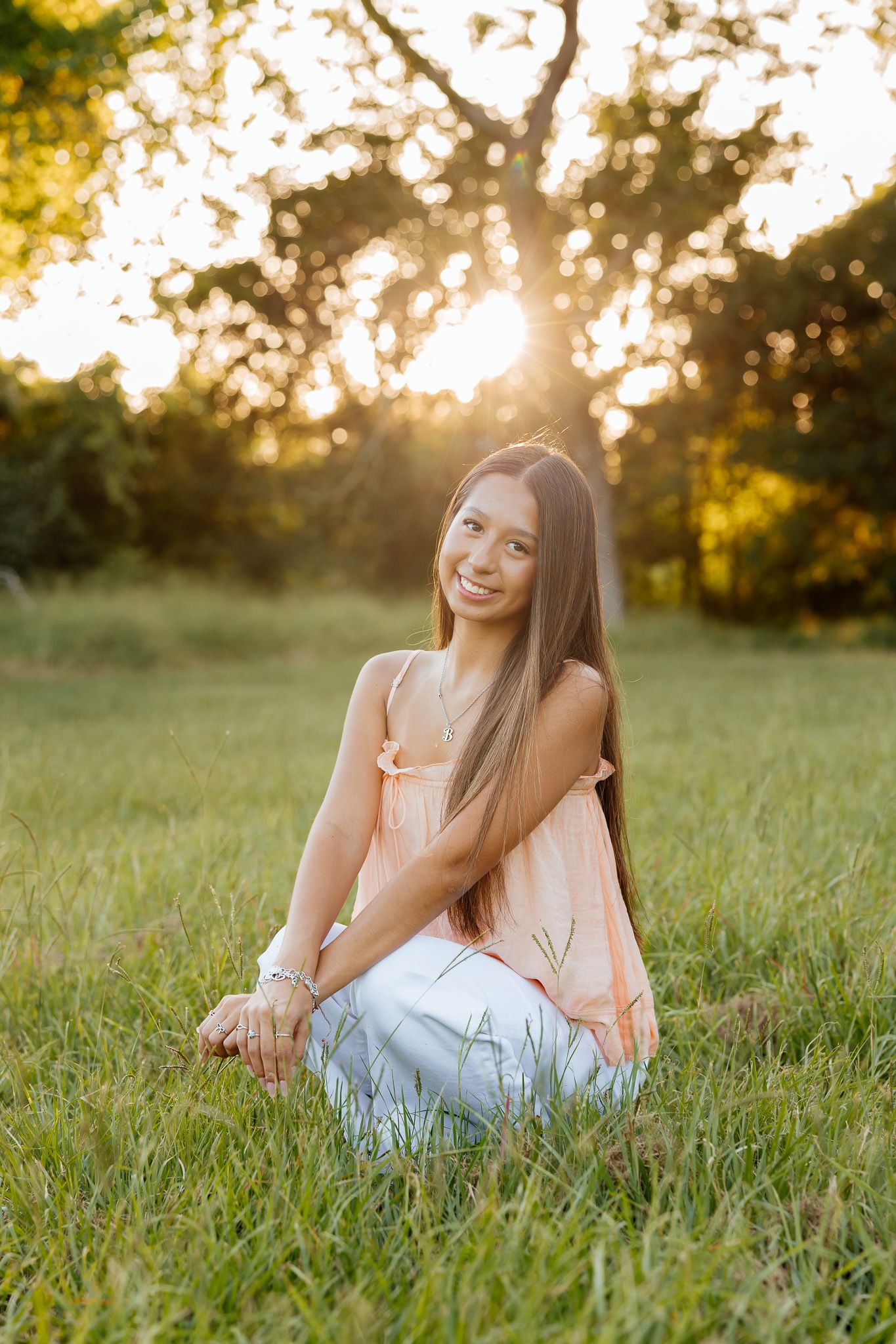 Young woman in pink top and white pants sitting on grass in golden sunset light, smiling outdoors.