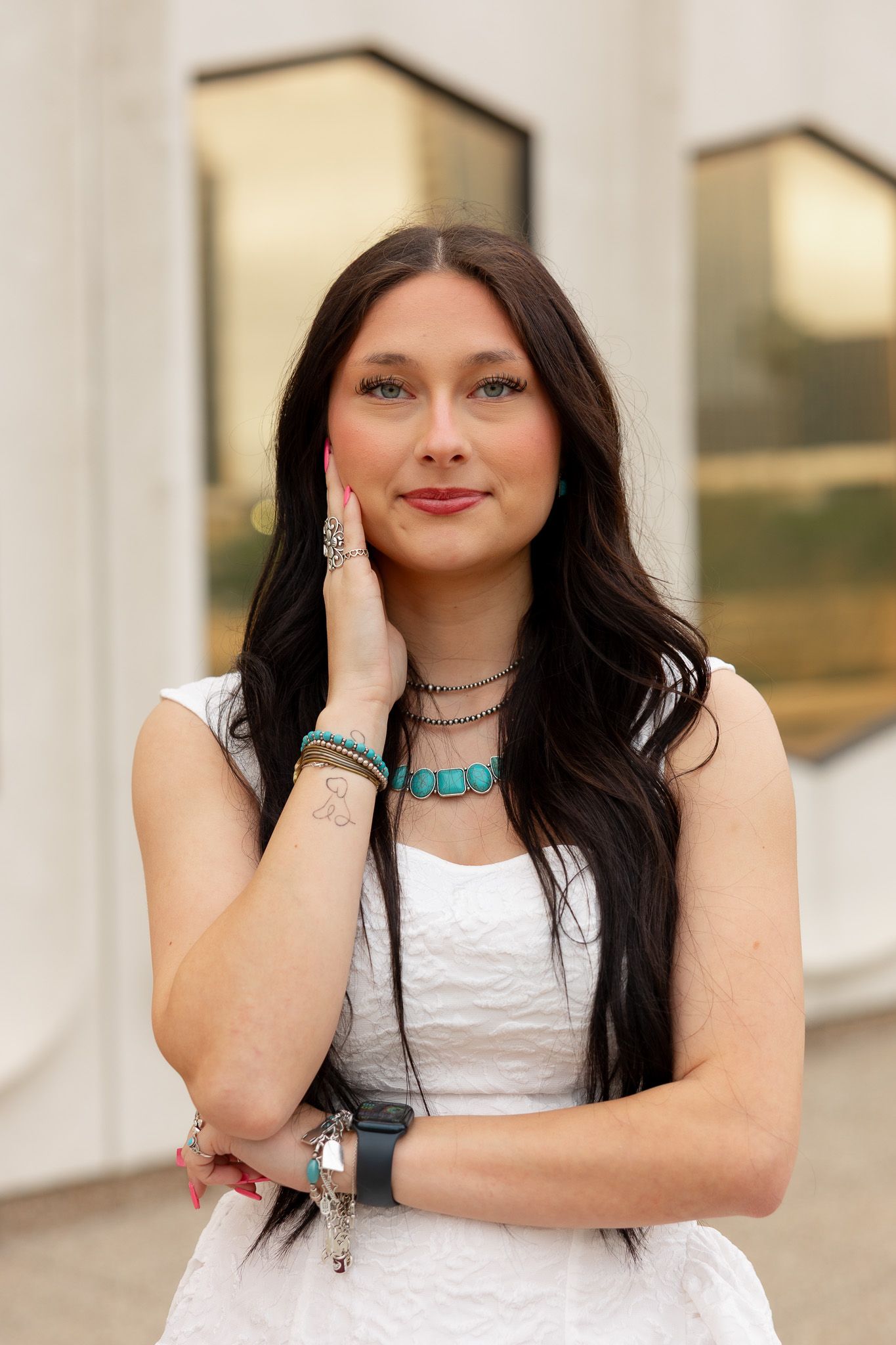 Young woman with long dark hair in white dress wearing turquoise jewelry poses outdoors.