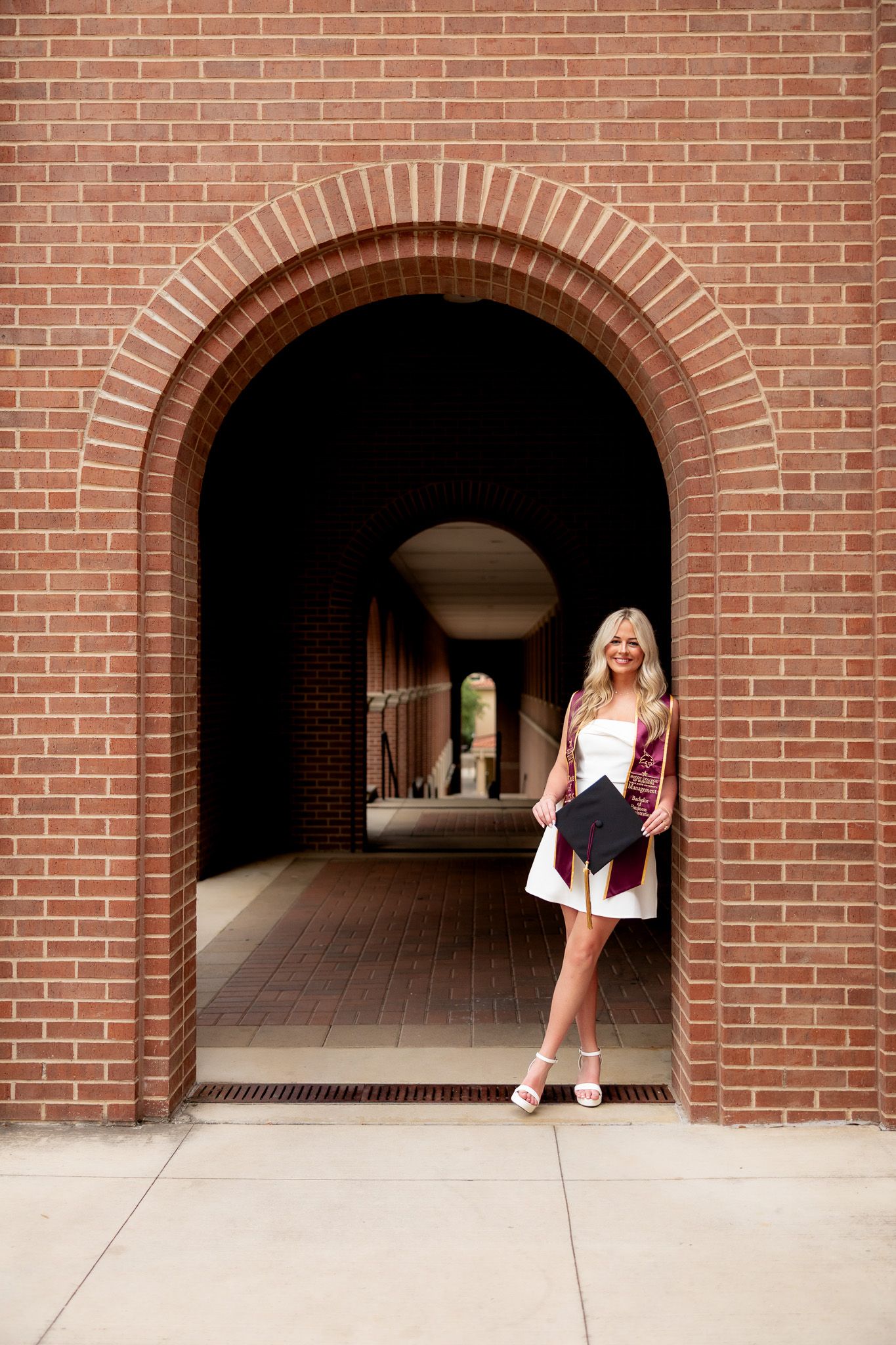 Blonde woman in white dress holding graduation cap posing in brick archway tunnel.