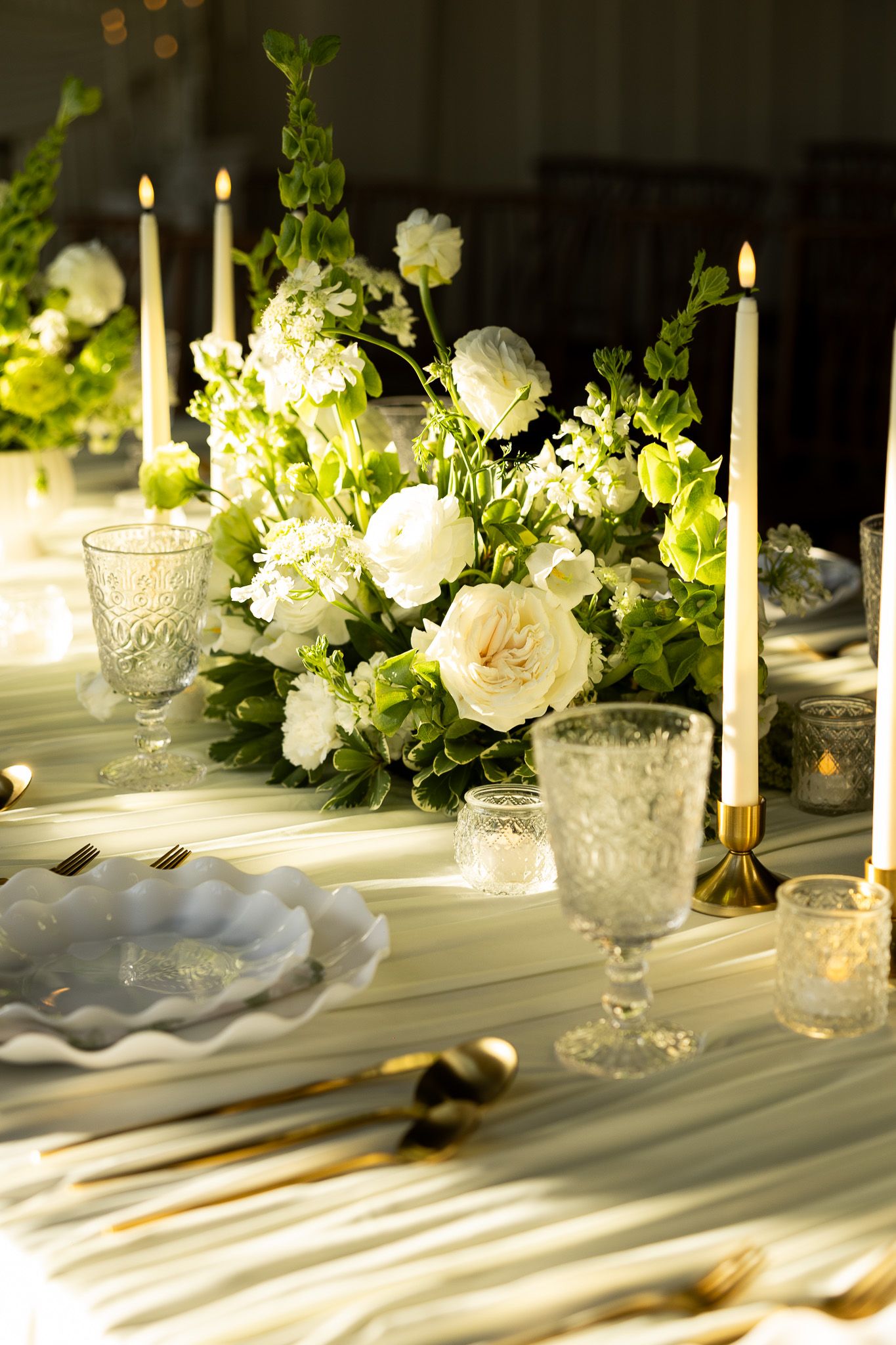 Elegant wedding table setting with white floral centerpiece, taper candles, crystal glasses, and gold cutlery on white linen.