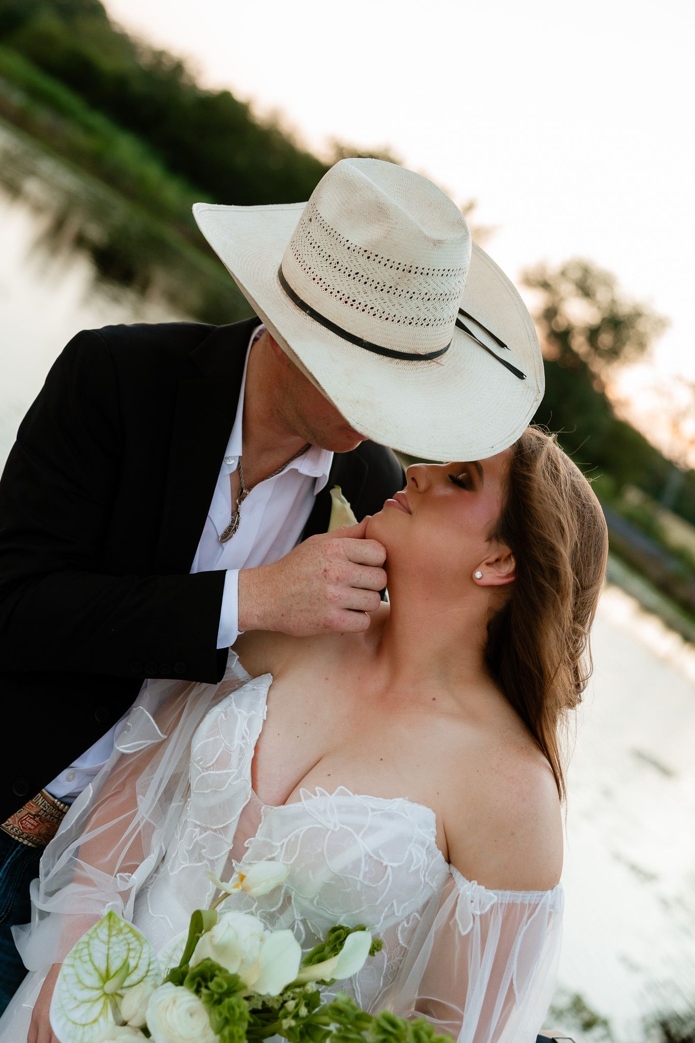 A groom in a white cowboy hat and black suit kisses his bride in a white lace dress outdoors near a lake.