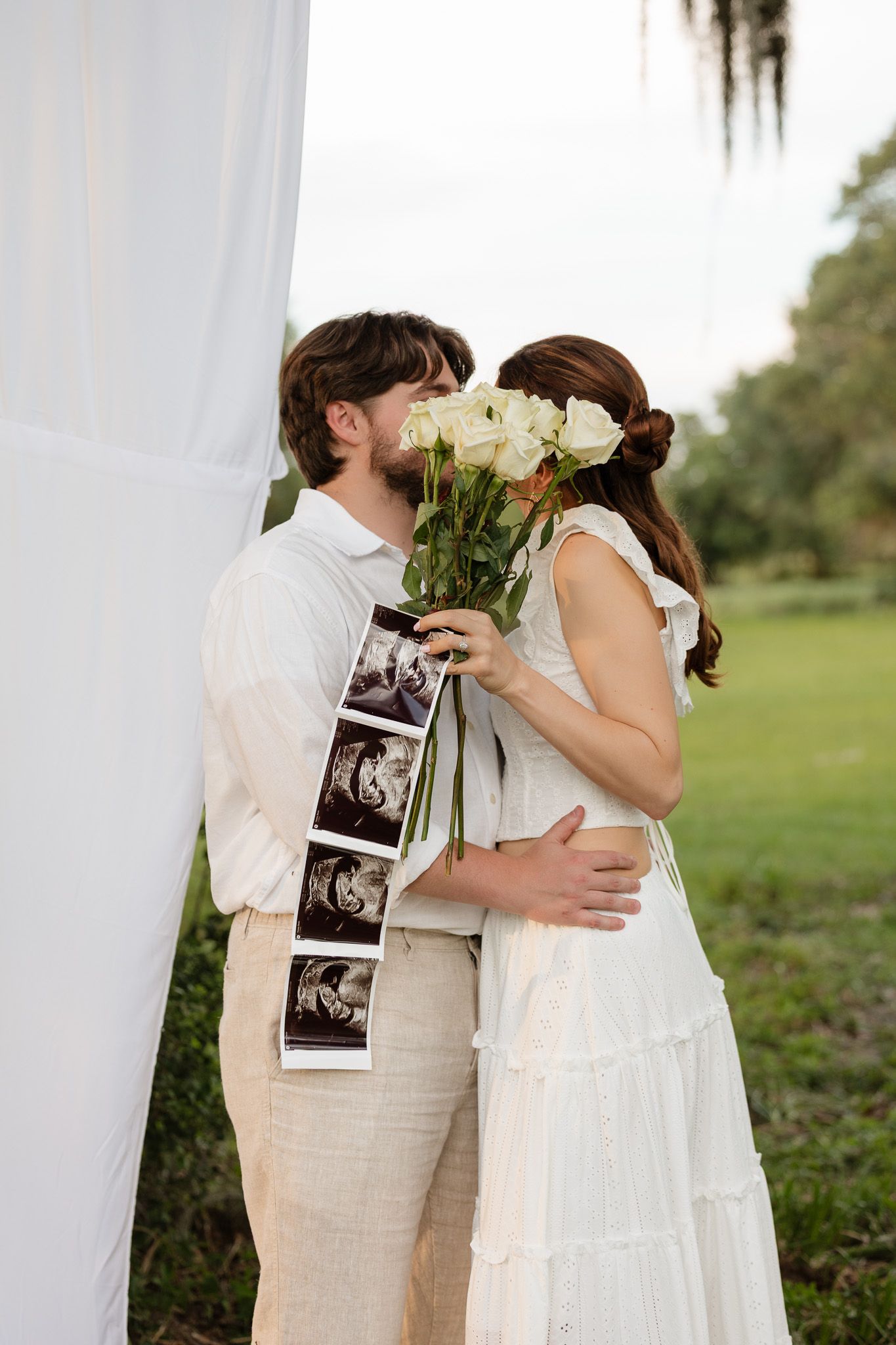Couple kissing behind white roses bouquet with ultrasound photos, wearing white outfits at outdoor wedding ceremony.
