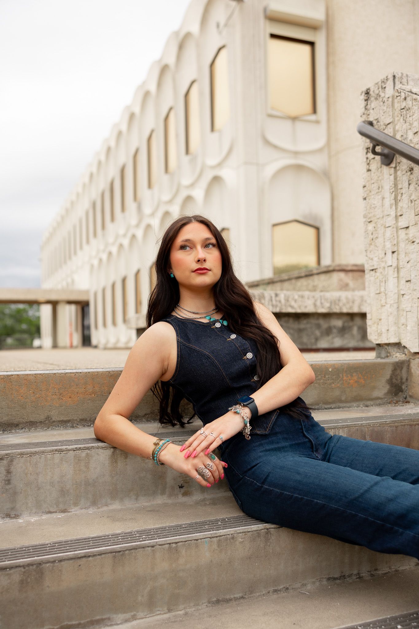 Young woman with long dark hair sits on stone steps wearing jeans, dark top, turquoise jewelry, posing outdoors.