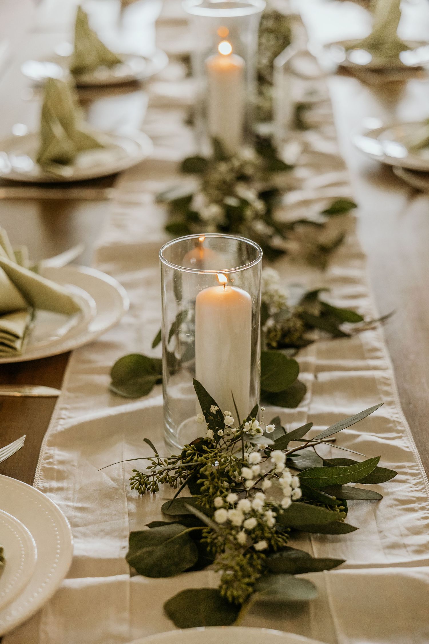 Elegant wedding reception table with white candles in glass holders and greenery centerpieces.
