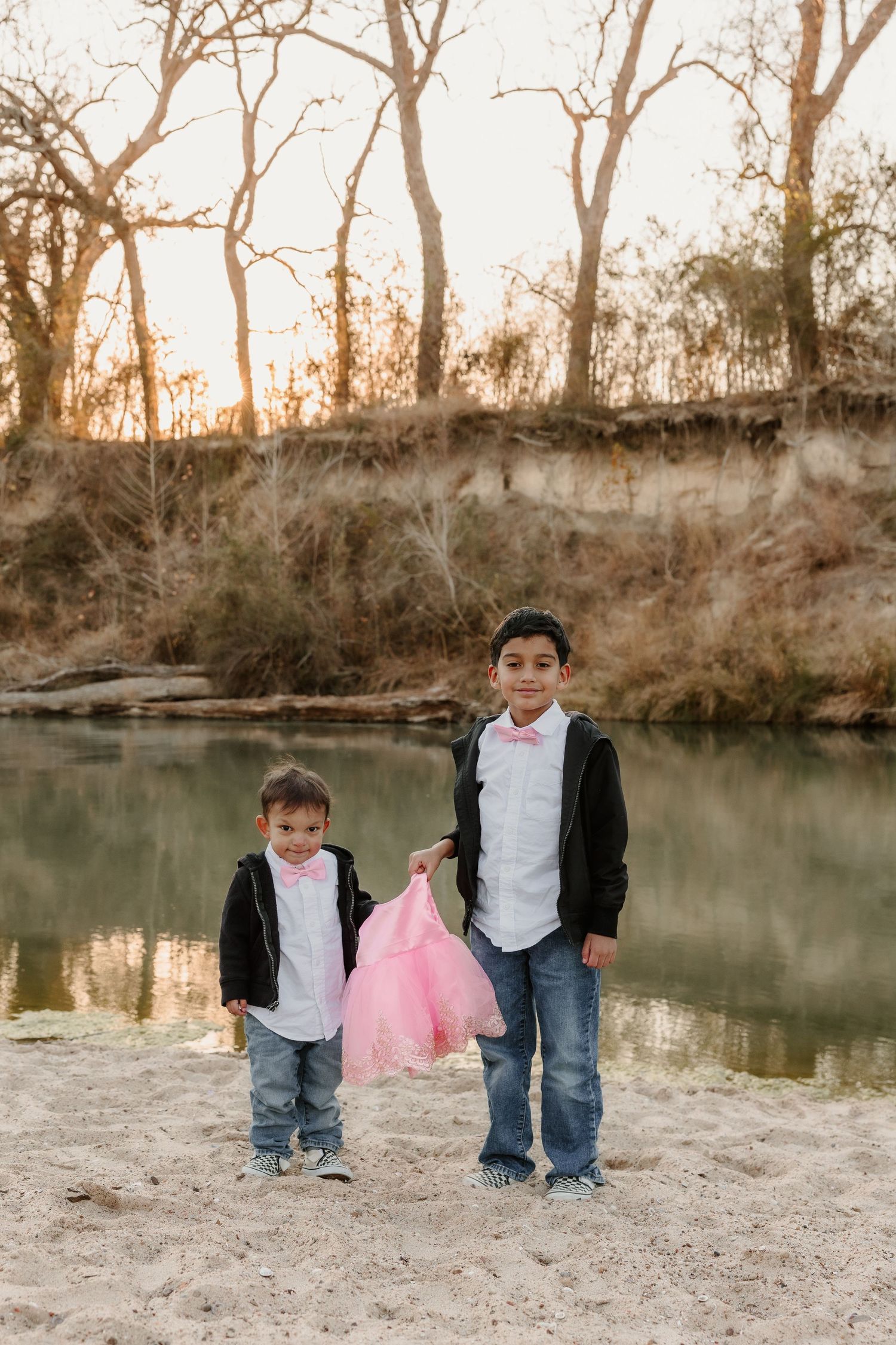 Two young boys in matching black jackets and pink bow ties stand on a sandy riverbank at sunset.