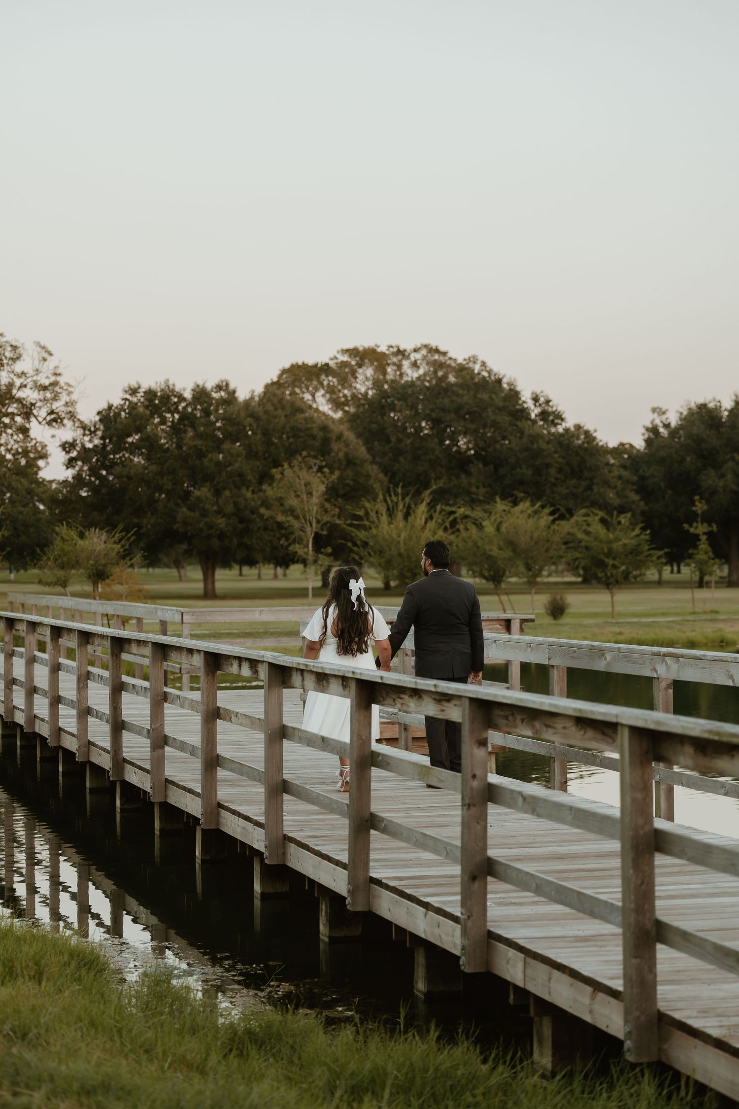 Couple walks hand in hand across long wooden boardwalk over water during sunset.