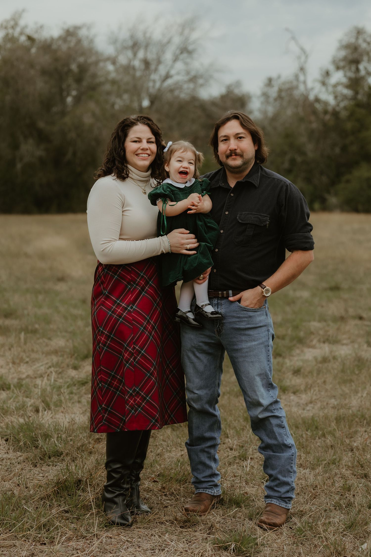 Family of three posing outdoors in a field, parents holding toddler in green dress, wearing holiday plaid and casual attire.