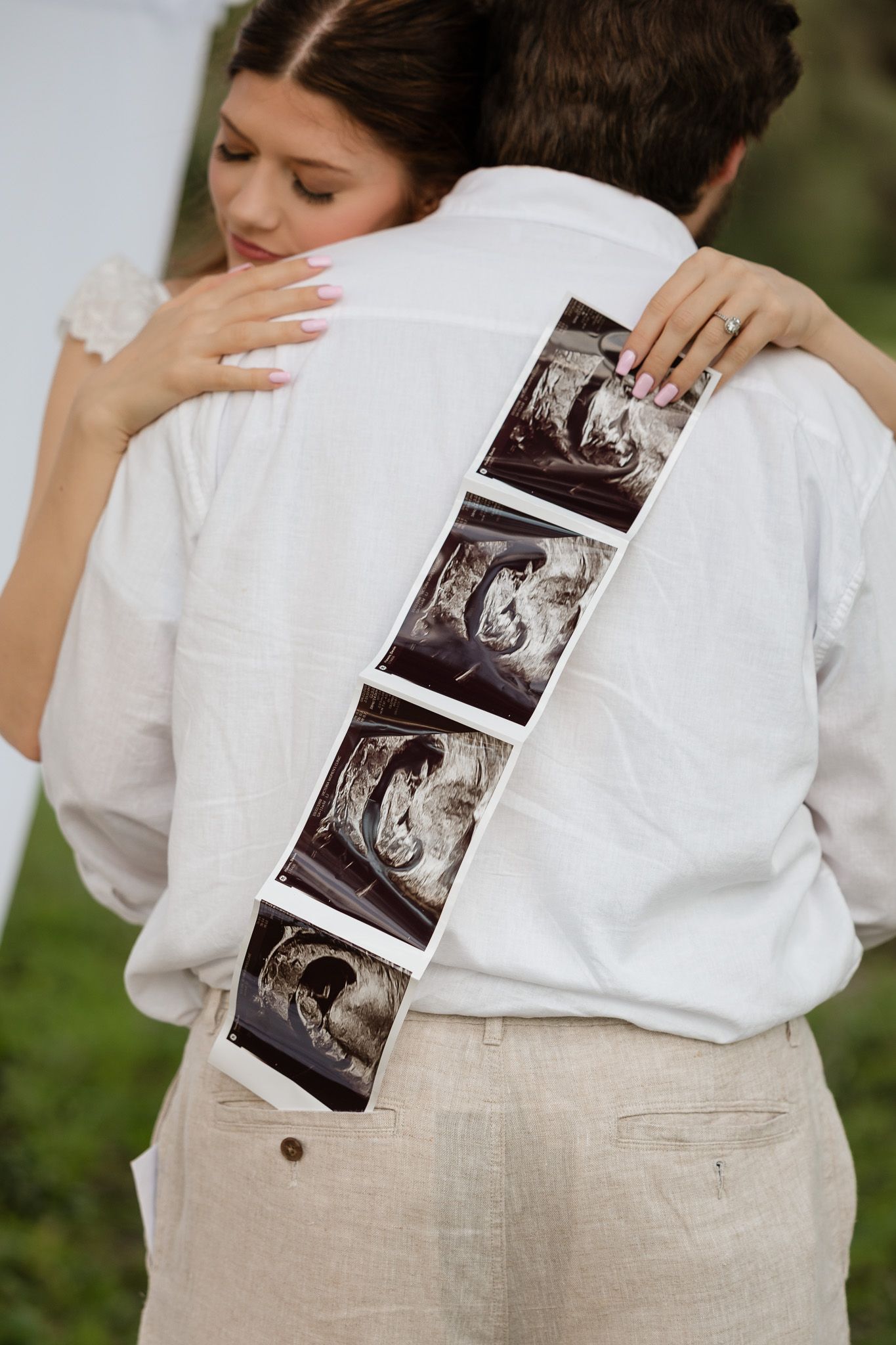 Couple embracing while holding ultrasound photos on man's back, announcing pregnancy at outdoor event.