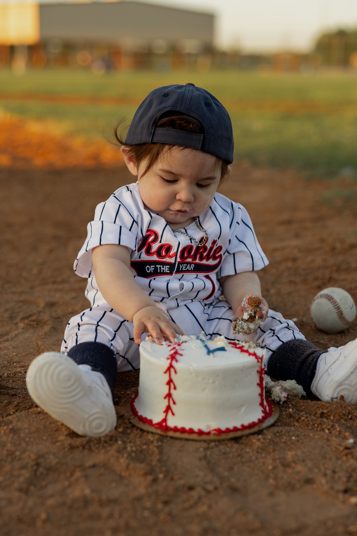 Baby in baseball uniform and cap touches a baseball-themed smash cake on a dirt field for a first birthday photo.