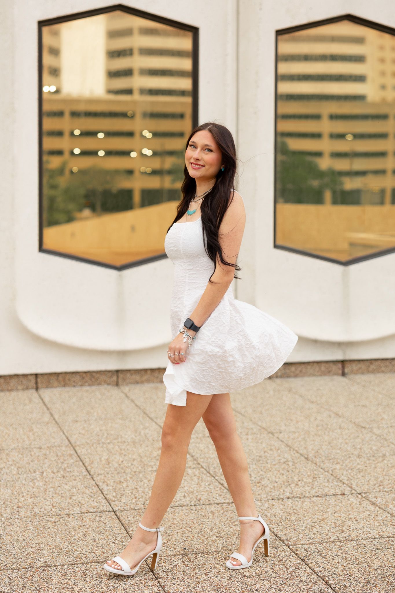 Young woman in white mini dress and heels walking outdoors near modern building windows.