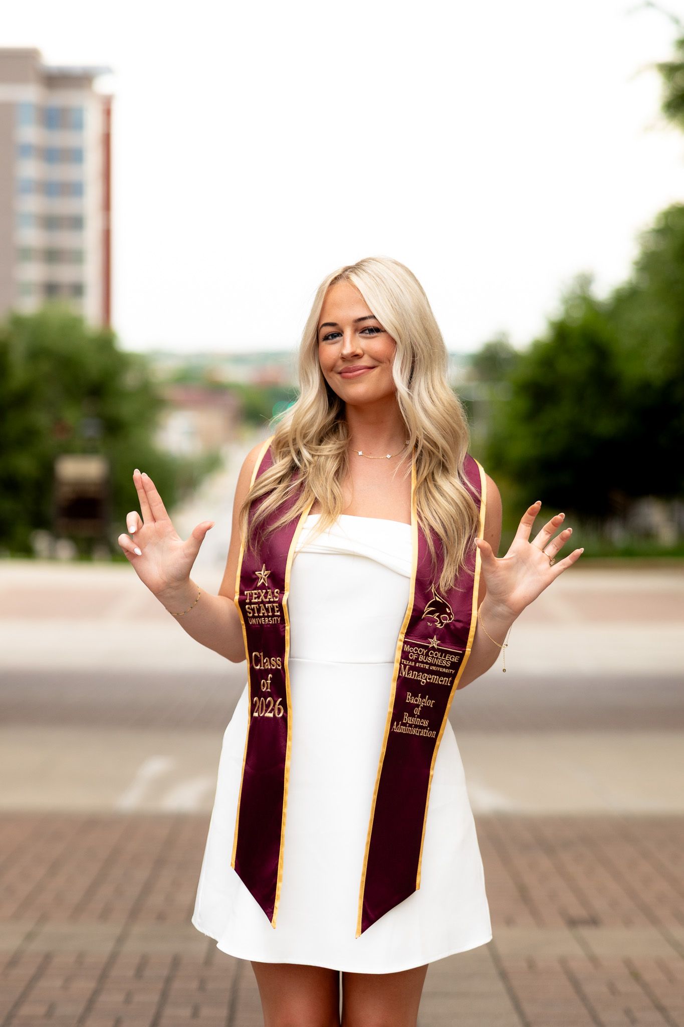 Smiling blonde woman in white dress wearing Texas State University Class of 2026 graduation stole outdoors.