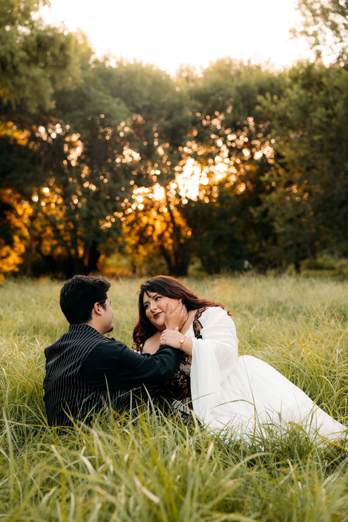 A couple sits in a lush green field at golden hour, sharing a tender moment with trees glowing in the sunset.