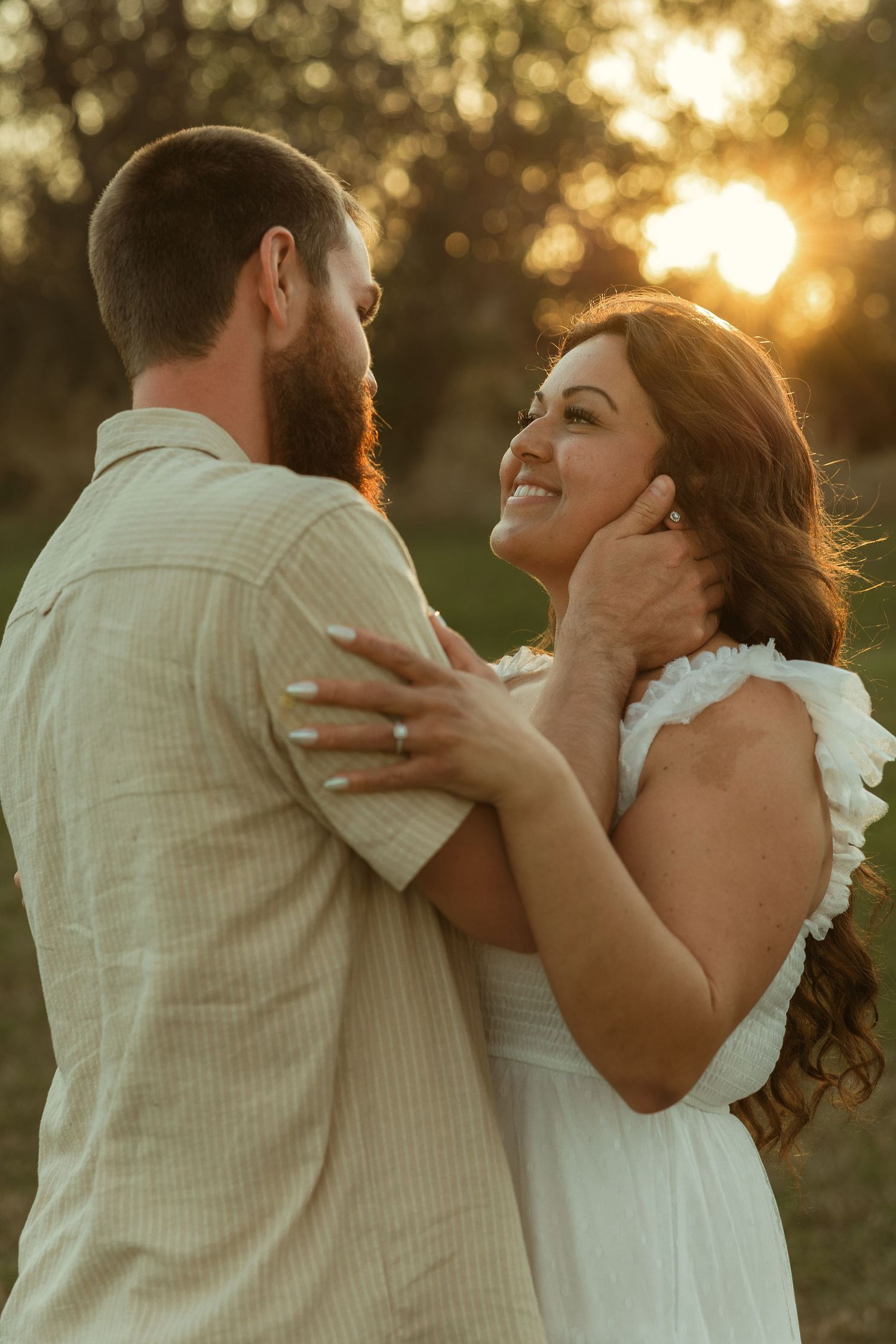 A smiling bride in a white dress gazes at her bearded groom during a golden sunset outdoor wedding photo.