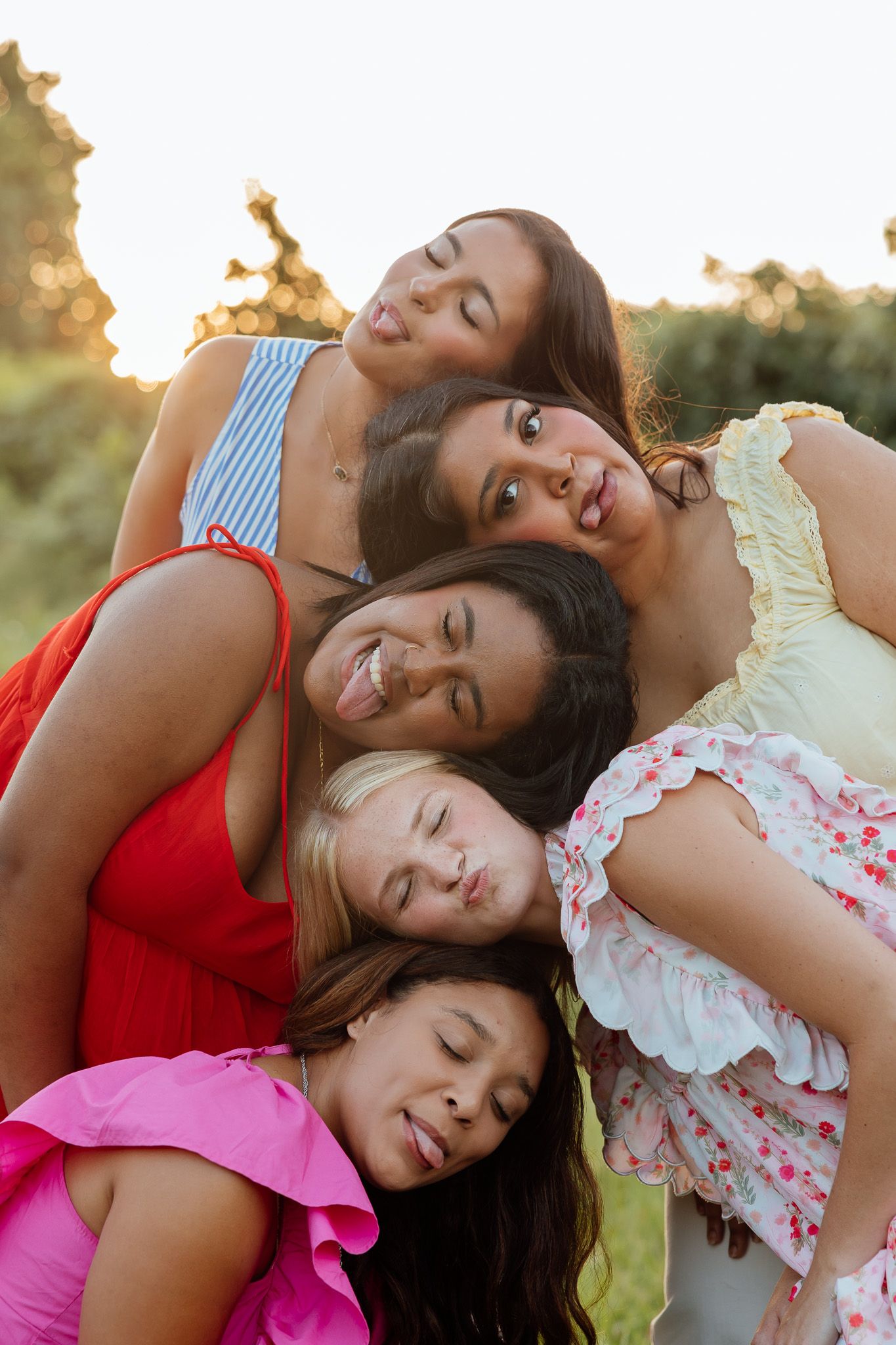 Five young women leaning heads together outdoors at golden hour, smiling and laughing in colorful summer dresses.