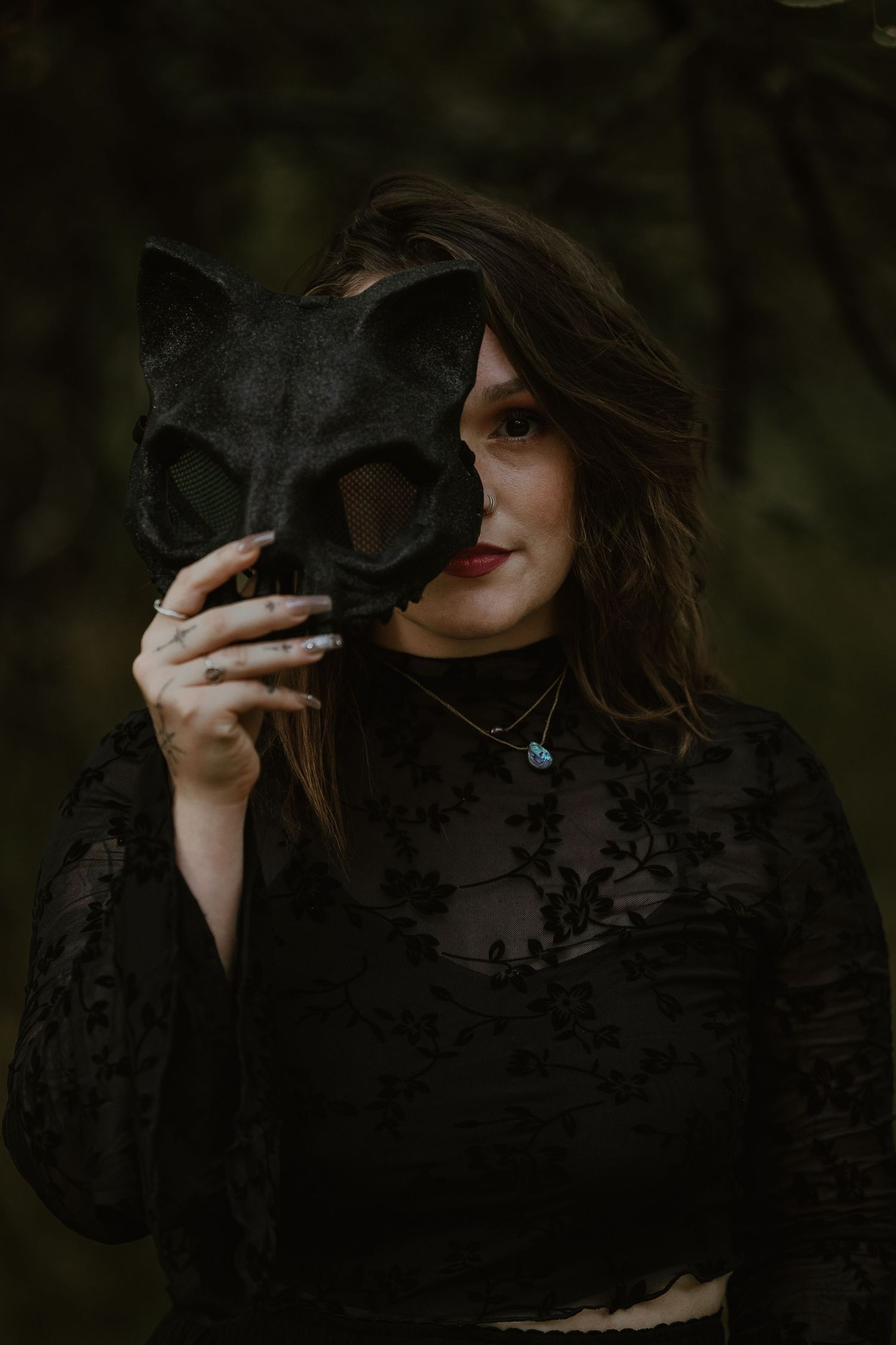 A moody portrait featuring a black cat mask and lace dress against a dark forest background.