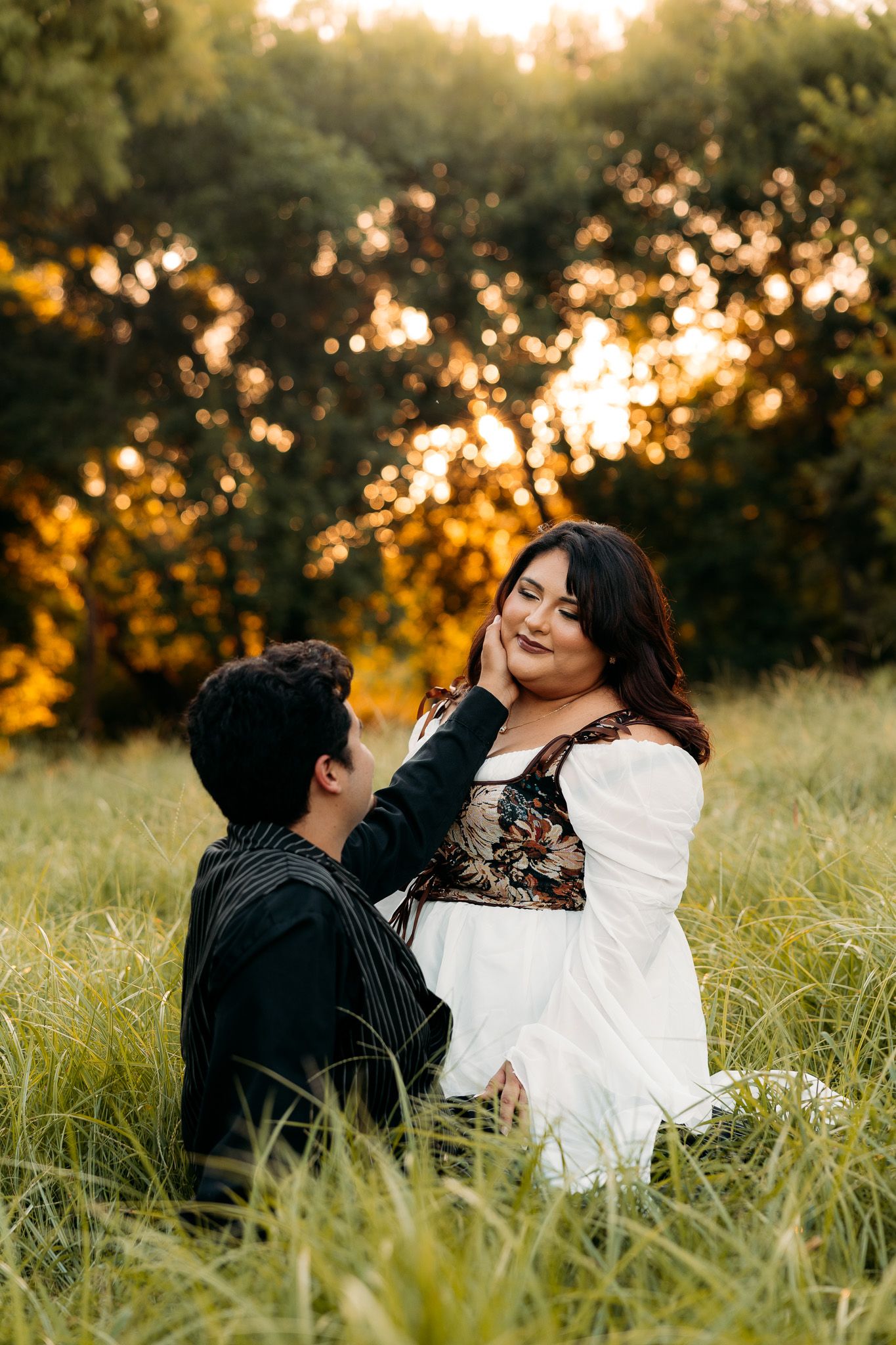 Couple sitting in a golden sunset meadow, woman in white dress tenderly touching man's face outdoors.