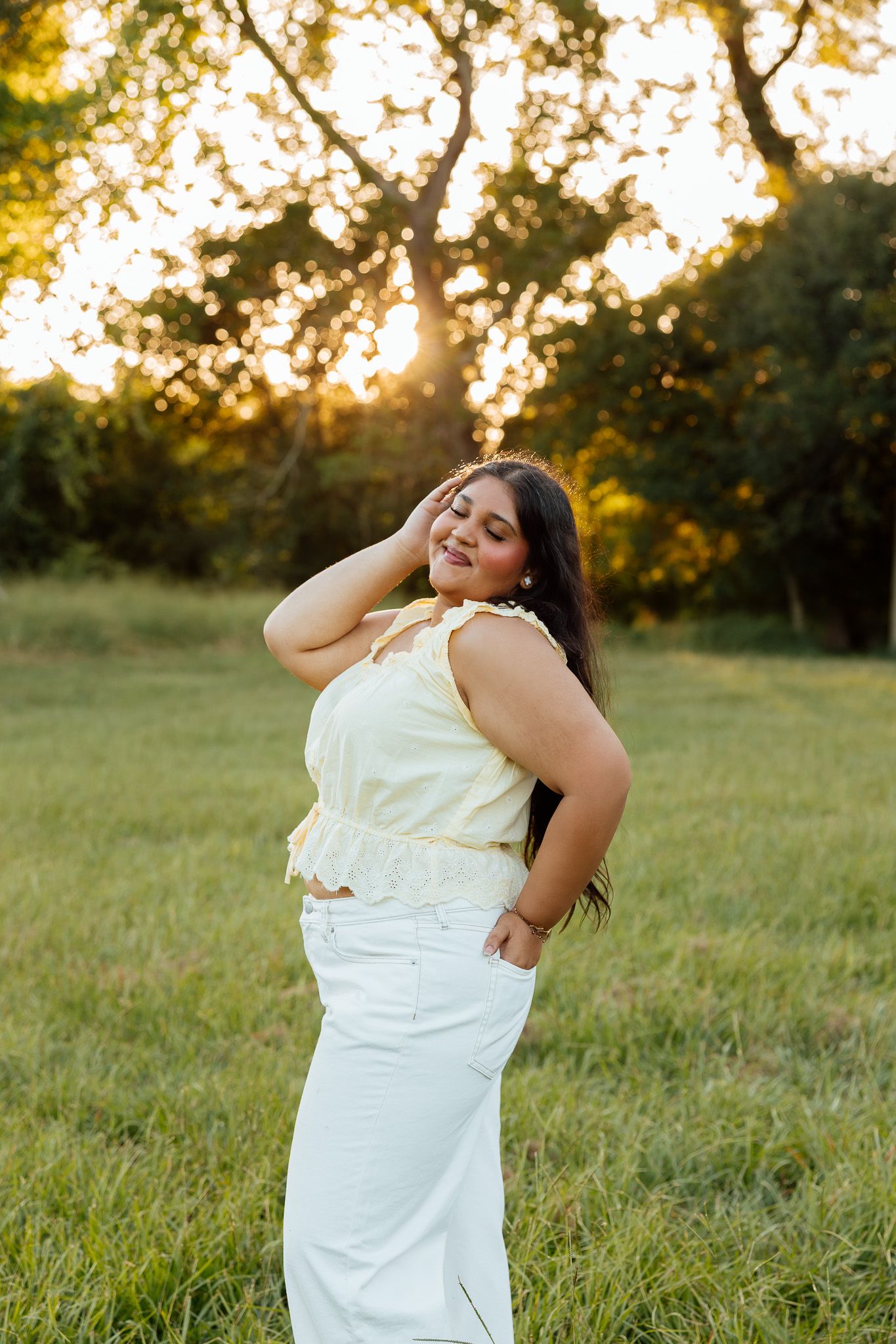 Young woman in yellow top and white skirt poses gracefully in a sunlit green field at golden hour.