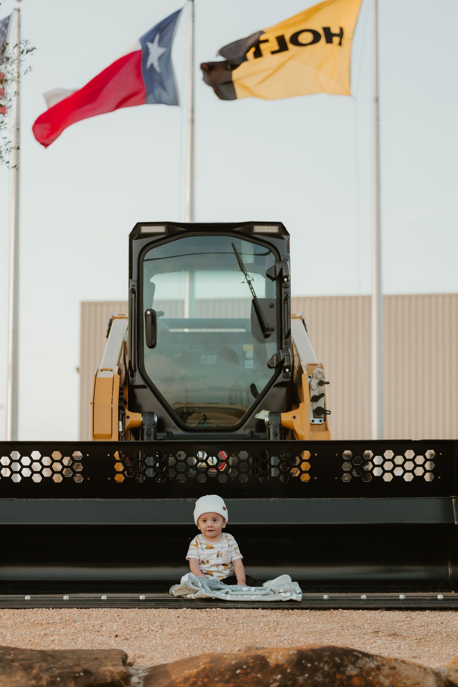 Baby sits before a yellow CAT skid steer loader at a Holt CAT dealership with Texas and company flags flying above.