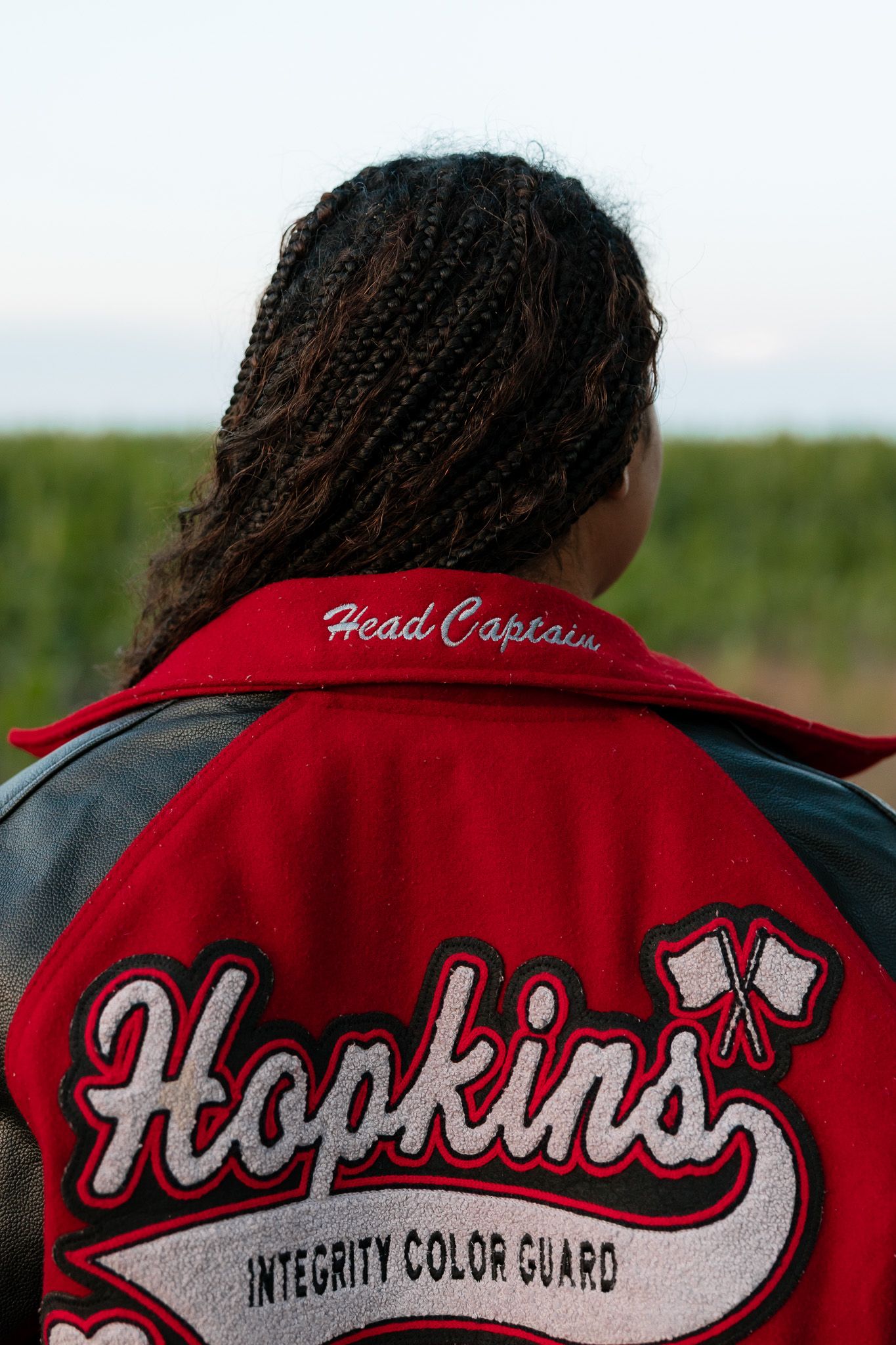 Person wearing a red Hopkins Integrity Color Guard Head Captain varsity jacket, standing outdoors facing away.