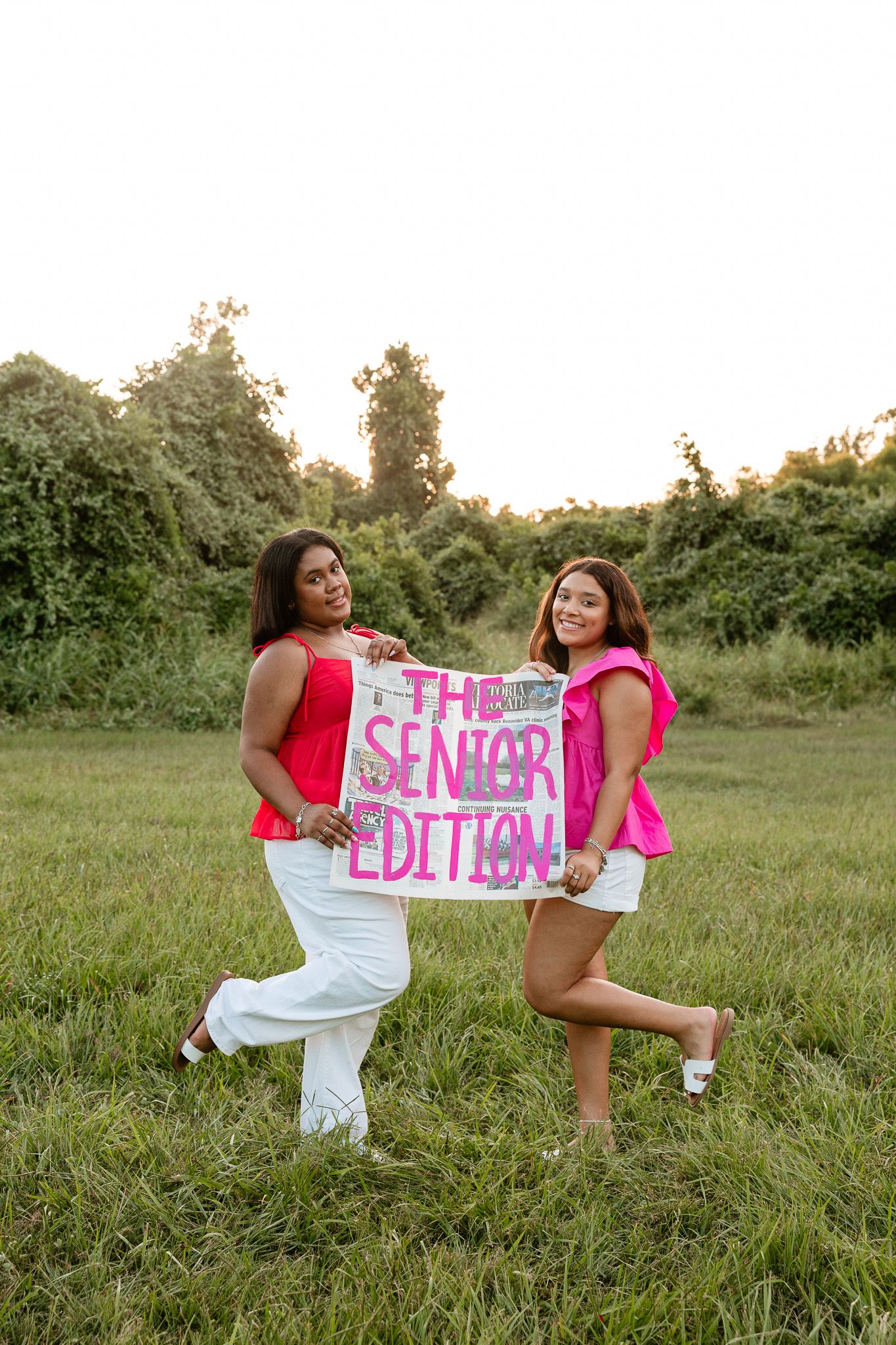 Two young women in pink tops and white bottoms hold a 'The Senior Edition' sign outdoors in a green field.