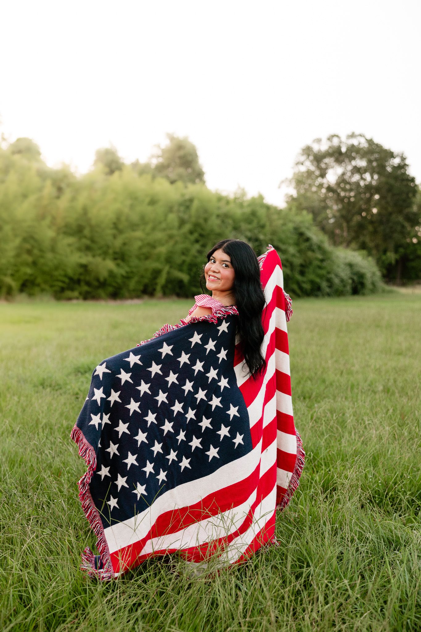 Young woman wrapped in an American flag blanket standing in a green field, smiling outdoors.