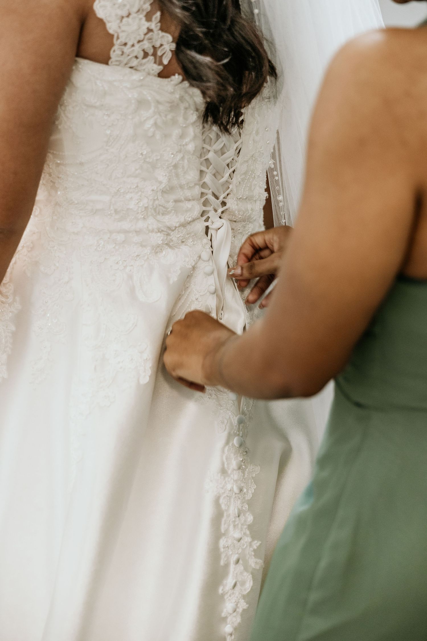 Close up of a white lace wedding dress being laced up by someone in a mint green dress.