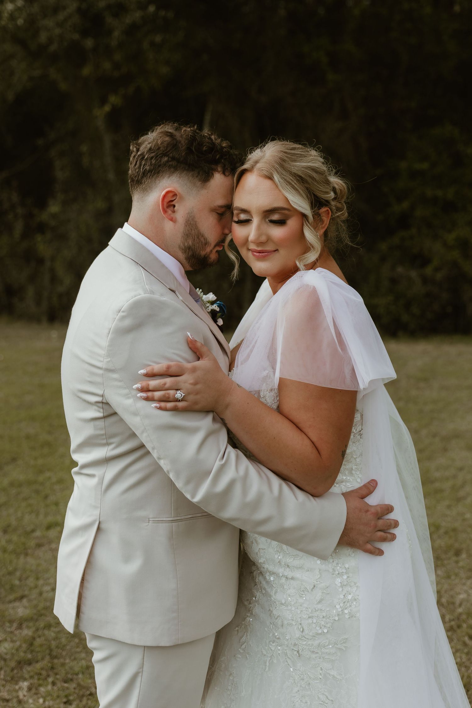 A romantic wedding couple embraces outdoors in a natural setting, wearing white attire and a flowing veil.