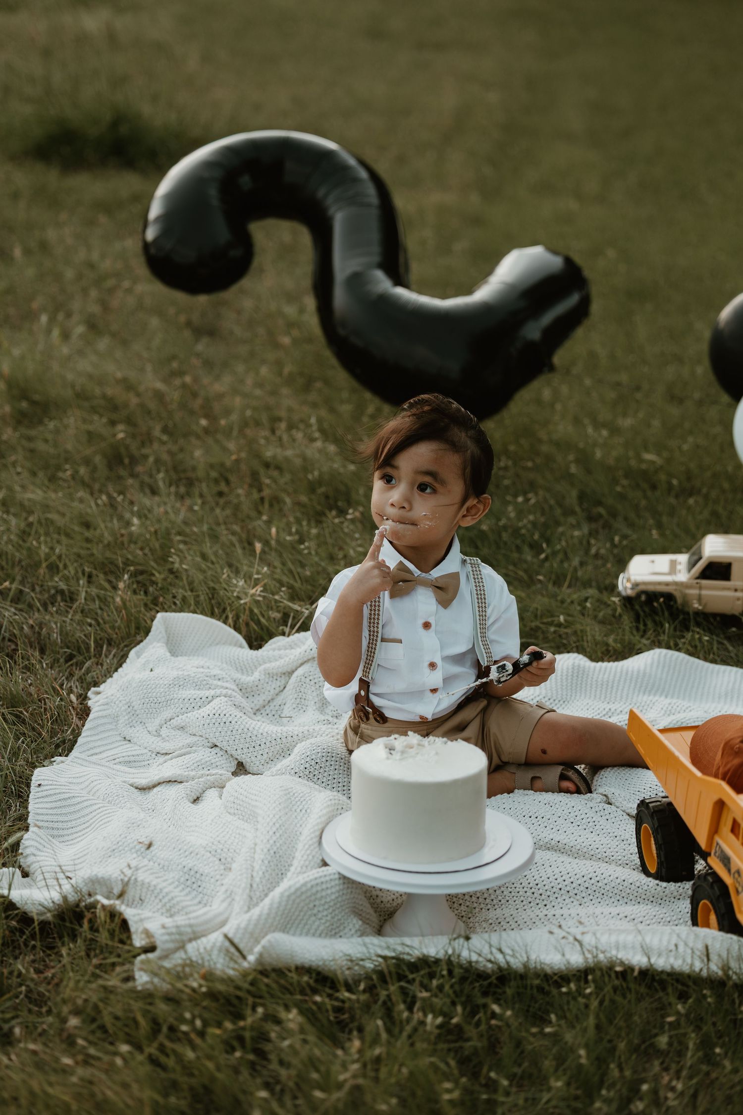 Child in formal wear sits on blanket with cake and toy truck under question mark balloon.