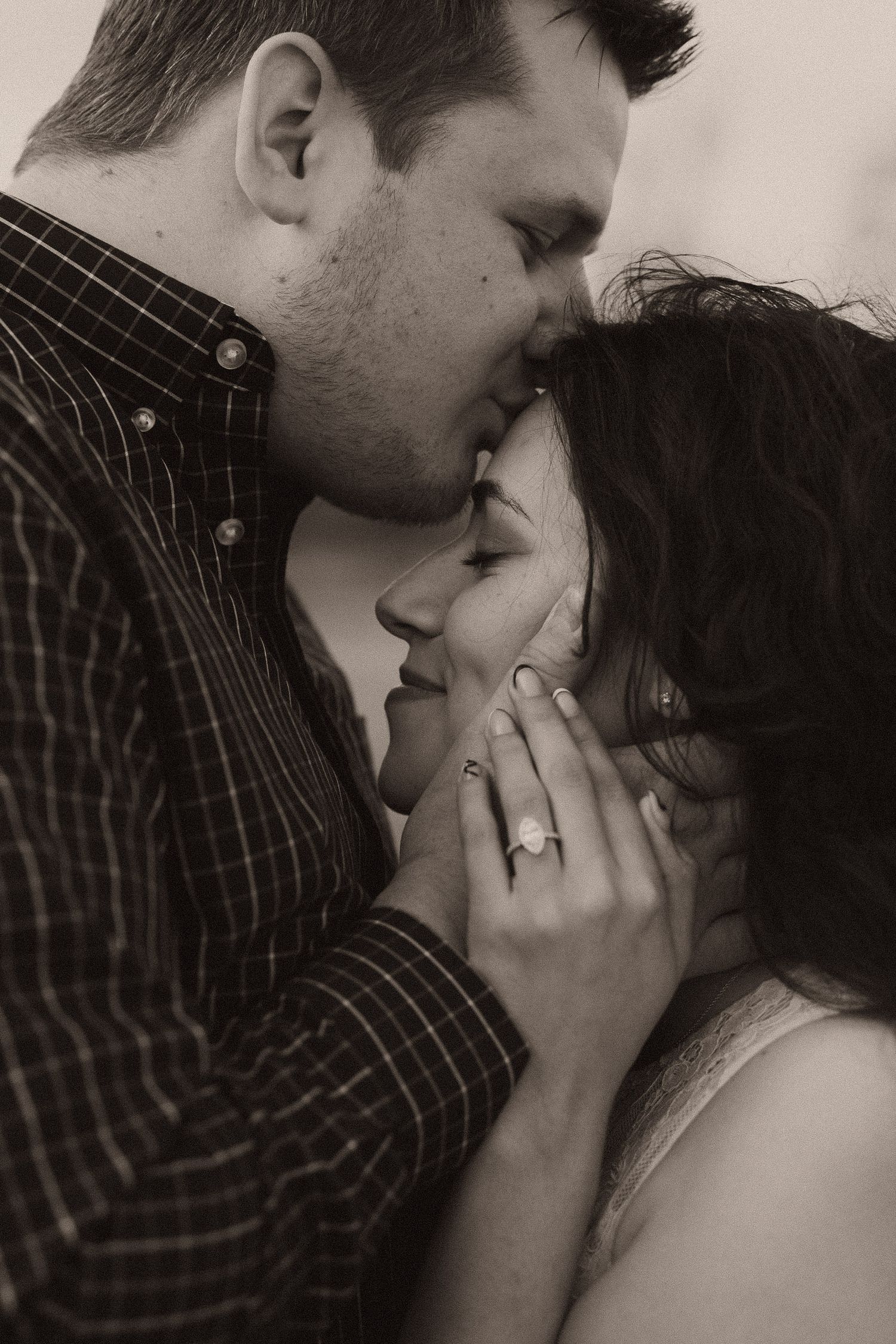 Black and white photo of a couple sharing a romantic moment, man tenderly holding womans face as they kiss, engagement ring visible.