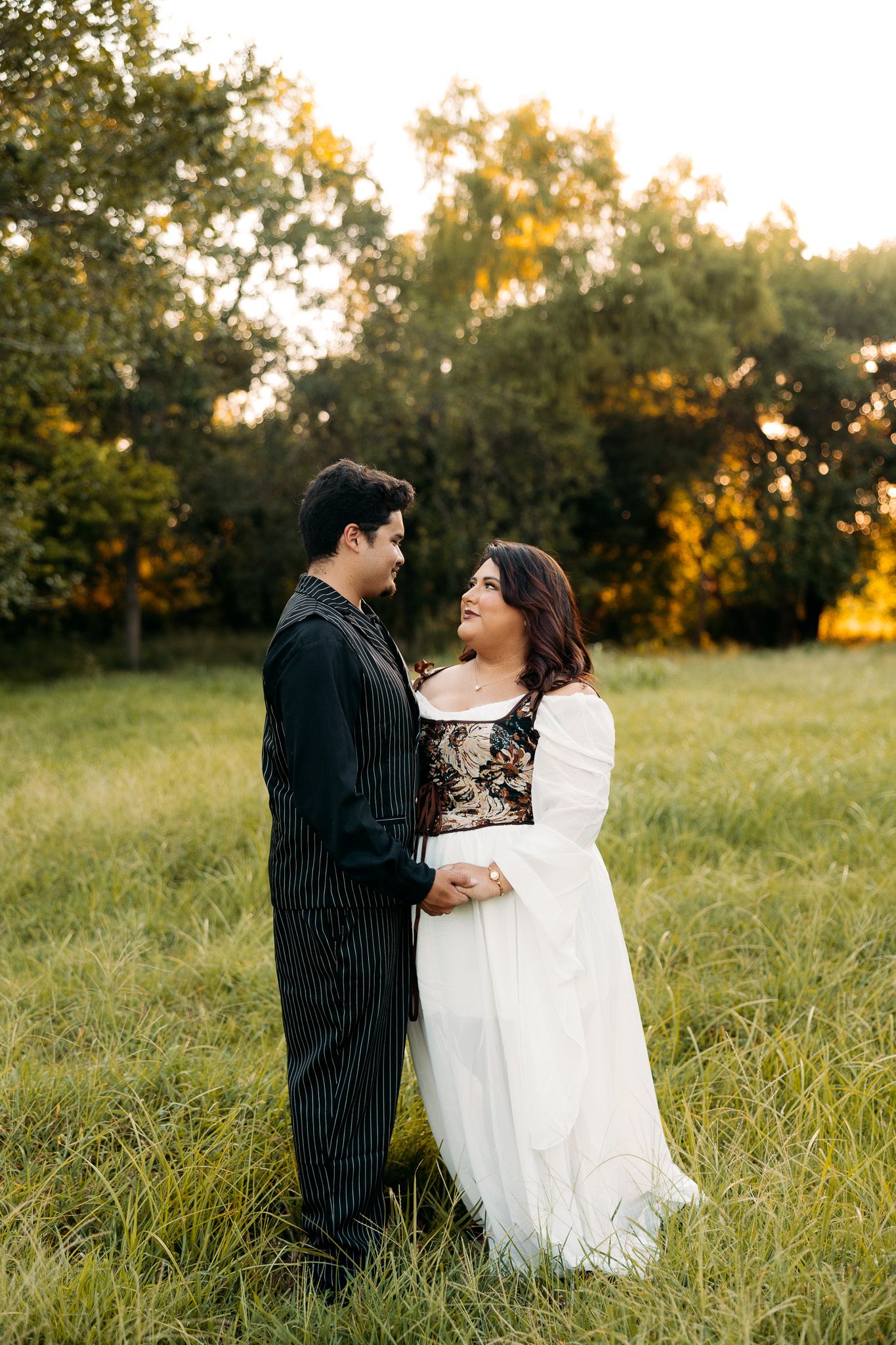 Couple in formal attire embracing in a lush green field at golden hour sunset.